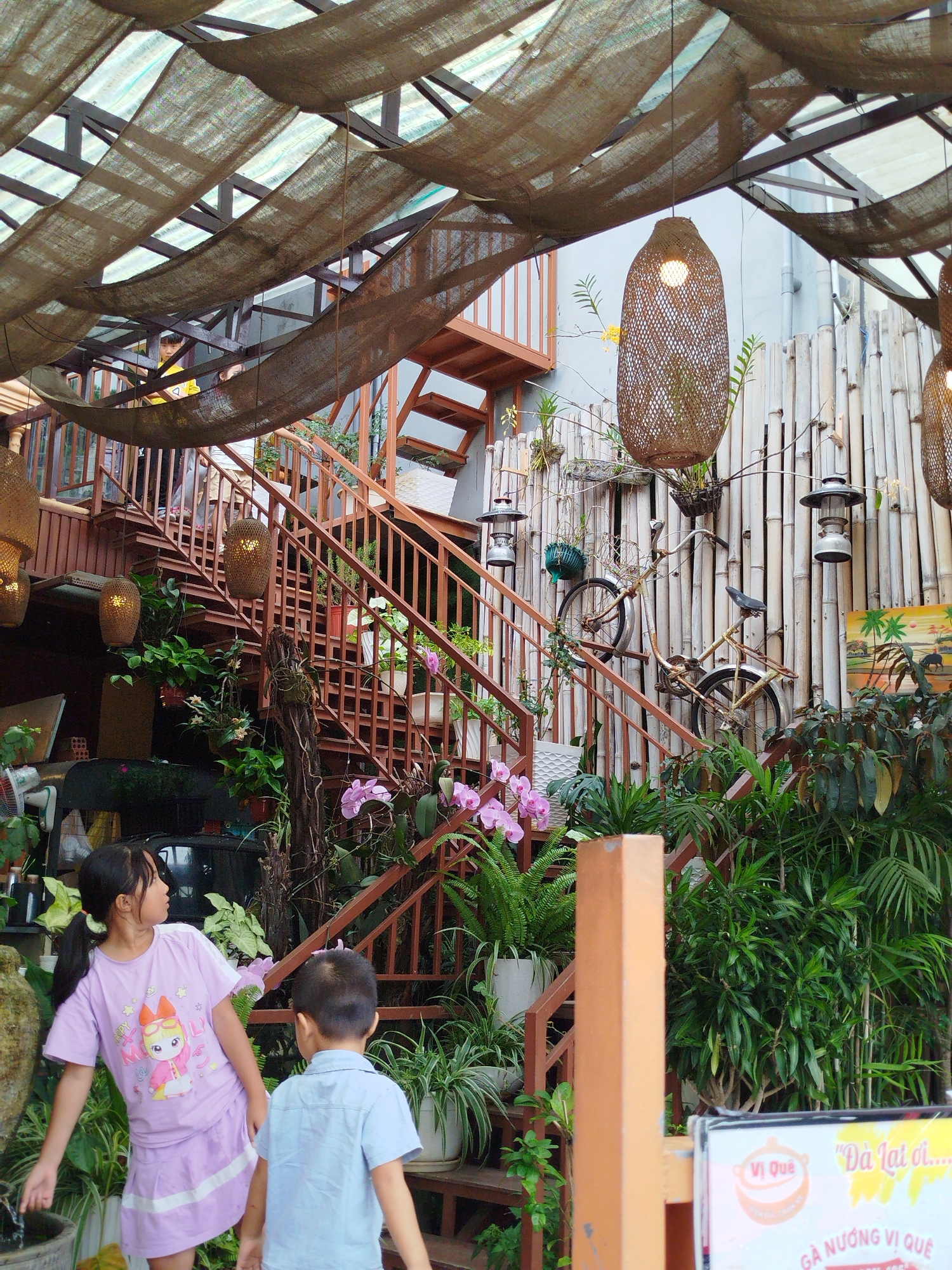 The image depicts an outdoor cafe or restaurant setting in Da Lat, Vietnam. The foreground features a young girl in a purple dress and a young boy in a blue shirt, both facing away from the camera. The girl has her hair in a ponytail and is looking to the left, while the boy is looking to the right. Behind them, a multi-tiered wooden and metal staircase ascends, adorned with various potted plants, including orchids. A rustic, woven lantern hangs from above. The background showcases a wall constructed of vertical bamboo poles, with a vintage bicycle mounted on it. Further up, a second, similar woven lantern and a kerosene lamp are visible. The cafe's roof is covered with a textured, dark brown fabric stretched over a metal frame, allowing dappled sunlight to filter through. Lush greenery surrounds the entire scene, characteristic of Da Lat's temperate climate and popular aesthetic for cafes. A small sign in the lower right corner displays Vietnamese text and graphics, indicating it is a commercial establishment.