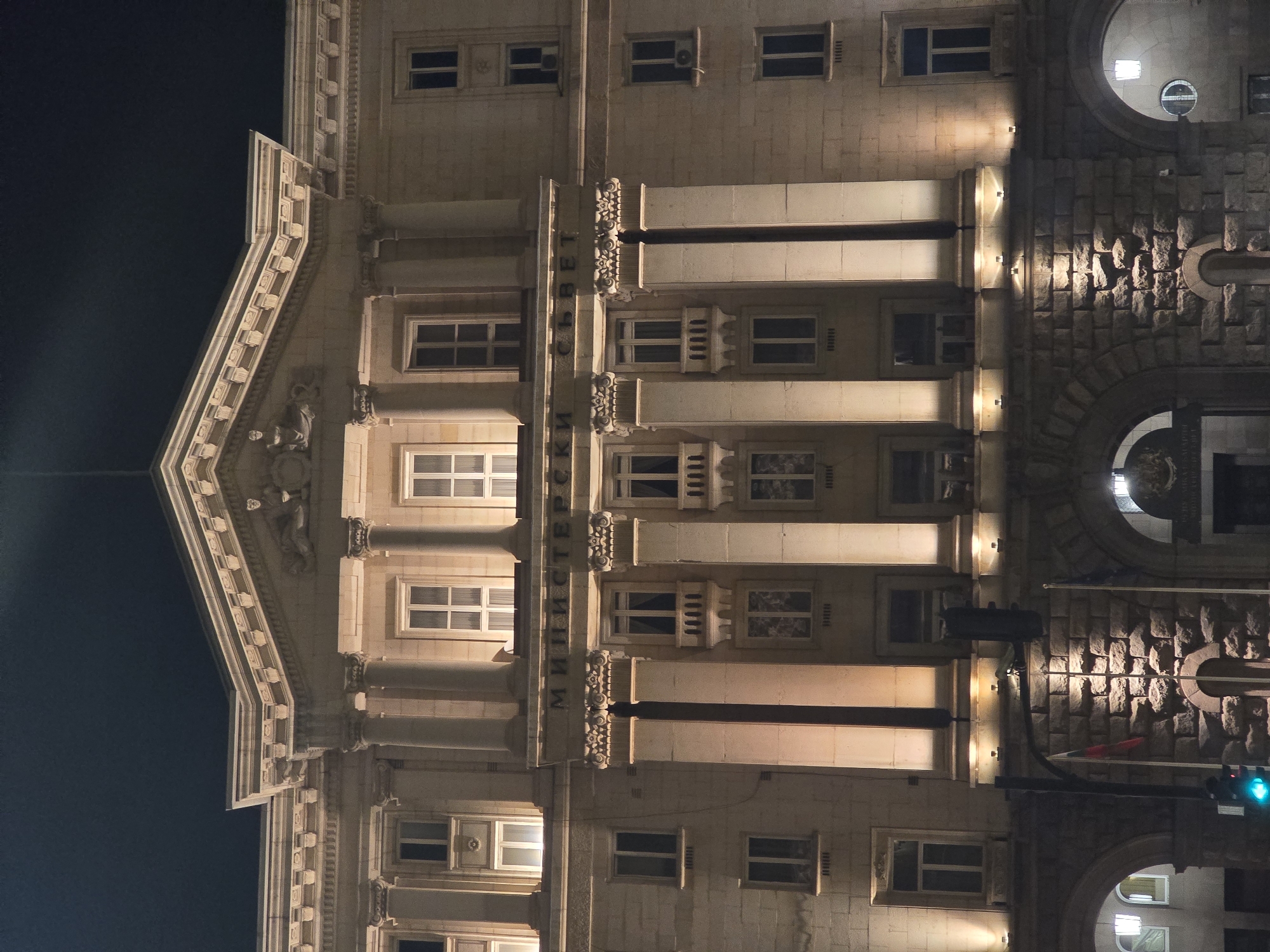 The image shows the facade of the Ministry of Justice building in Sofia, Bulgaria, at night. The building is made of light-colored stone and features a symmetrical design with classical architectural elements such as columns, windows, and a pediment. The facade is illuminated by artificial light, which emphasizes the details of the building's ornamentation. The building has a large arch. Some of the windows are lit from within. A traffic light is visible in front of the building. The sky is dark.