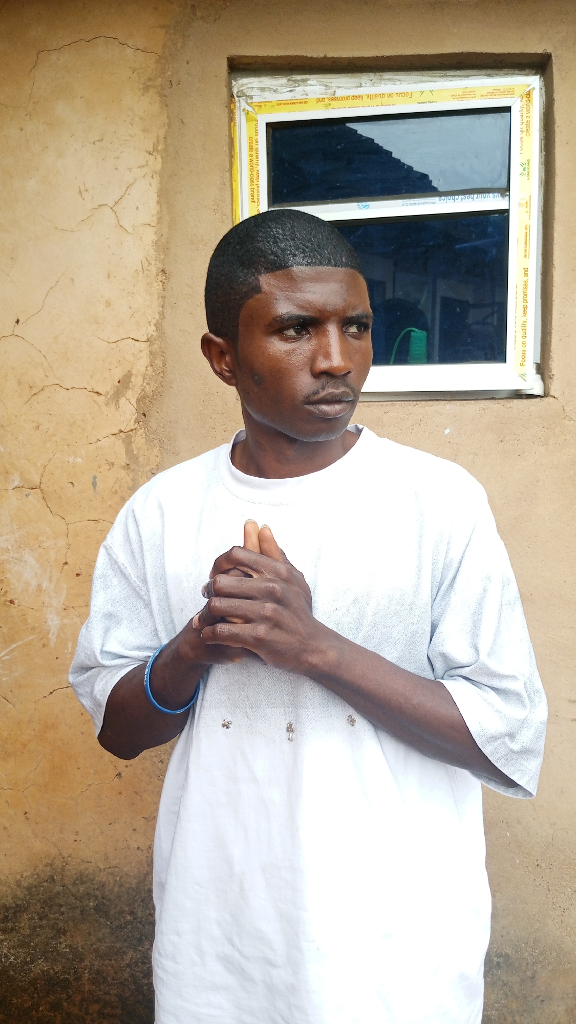 A young African man stands in front of a weathered, yellowish wall in Kuru Karama, Nigeria. A white-framed window with dark glass is visible behind him. The man is wearing a white t-shirt and a blue wristband. His hands are clasped together at chest level. The wall shows signs of age and cracking. The window frame has a yellow and white sticker with text.