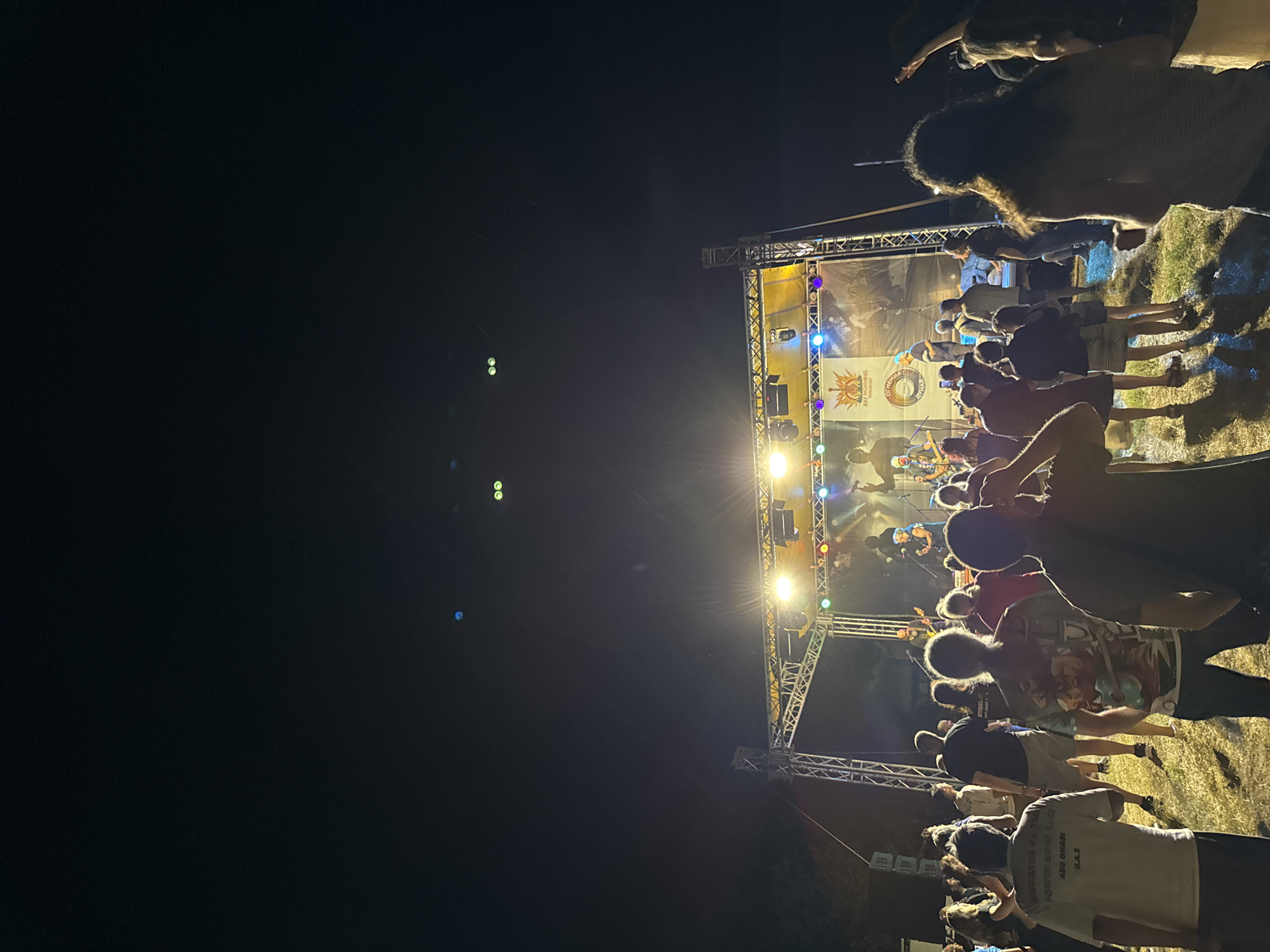 The image depicts an outdoor nighttime event in Tutrakan, Bulgaria, with a live band performing on a stage illuminated by bright lights. A crowd of people is gathered in front of the stage, some dancing and enjoying the music, while others are standing and observing. The stage is constructed from metal trussing and features a large banner at the back with text and graphics. The surrounding environment appears to be a grassy area, possibly a park or open field. In the distance, the night sky is visible, with a few faint lights or celestial bodies. The overall atmosphere suggests a concert or festival event.