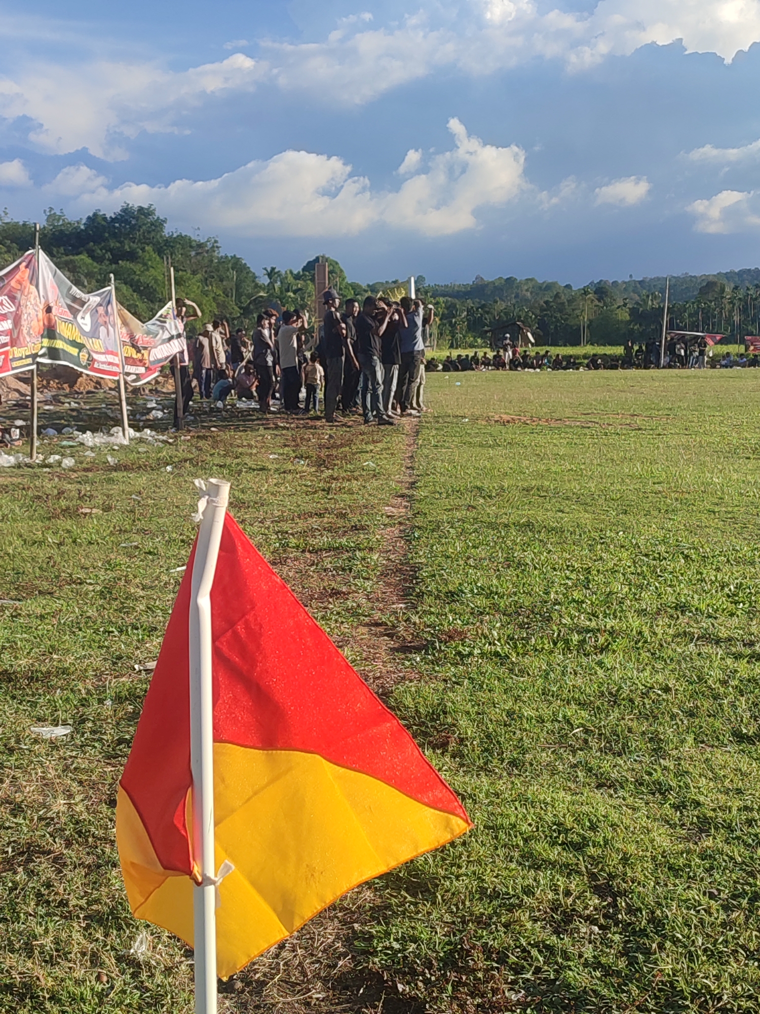 The image shows a grassy field on what appears to be a partly cloudy day in Babahlueng, Indonesia. In the foreground is a corner flag, red on the top half and yellow on the bottom half. In the background, a crowd of people is gathered near what seems to be a boundary, some standing, some seated. Banners are set up on poles on the left. The overall environment looks like a sporting event or community gathering.