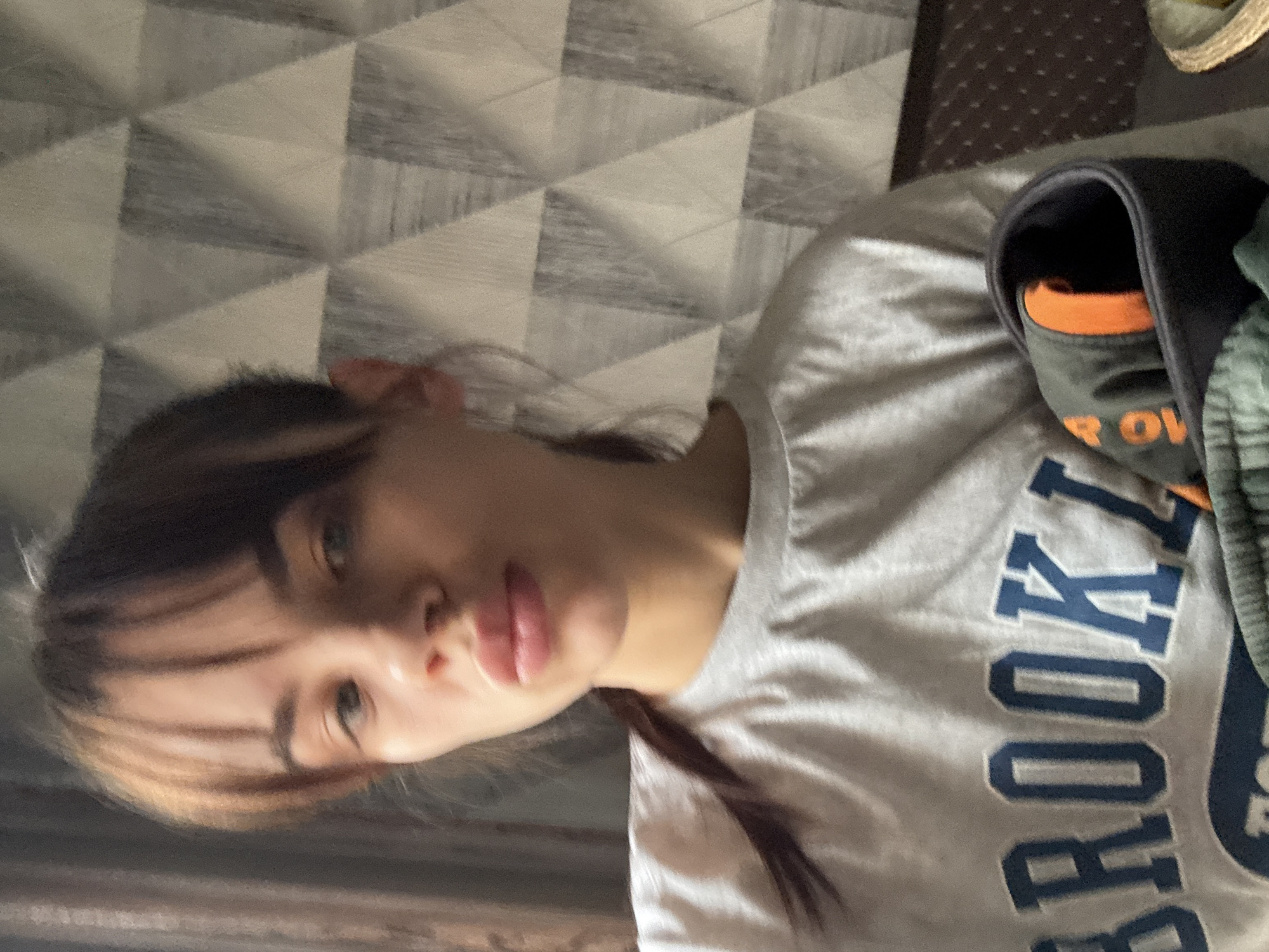 A woman is facing the camera, wearing a gray t-shirt with "BROOKLYN" printed on it in blue. The background is a geometric patterned wall. The location is not specified.
