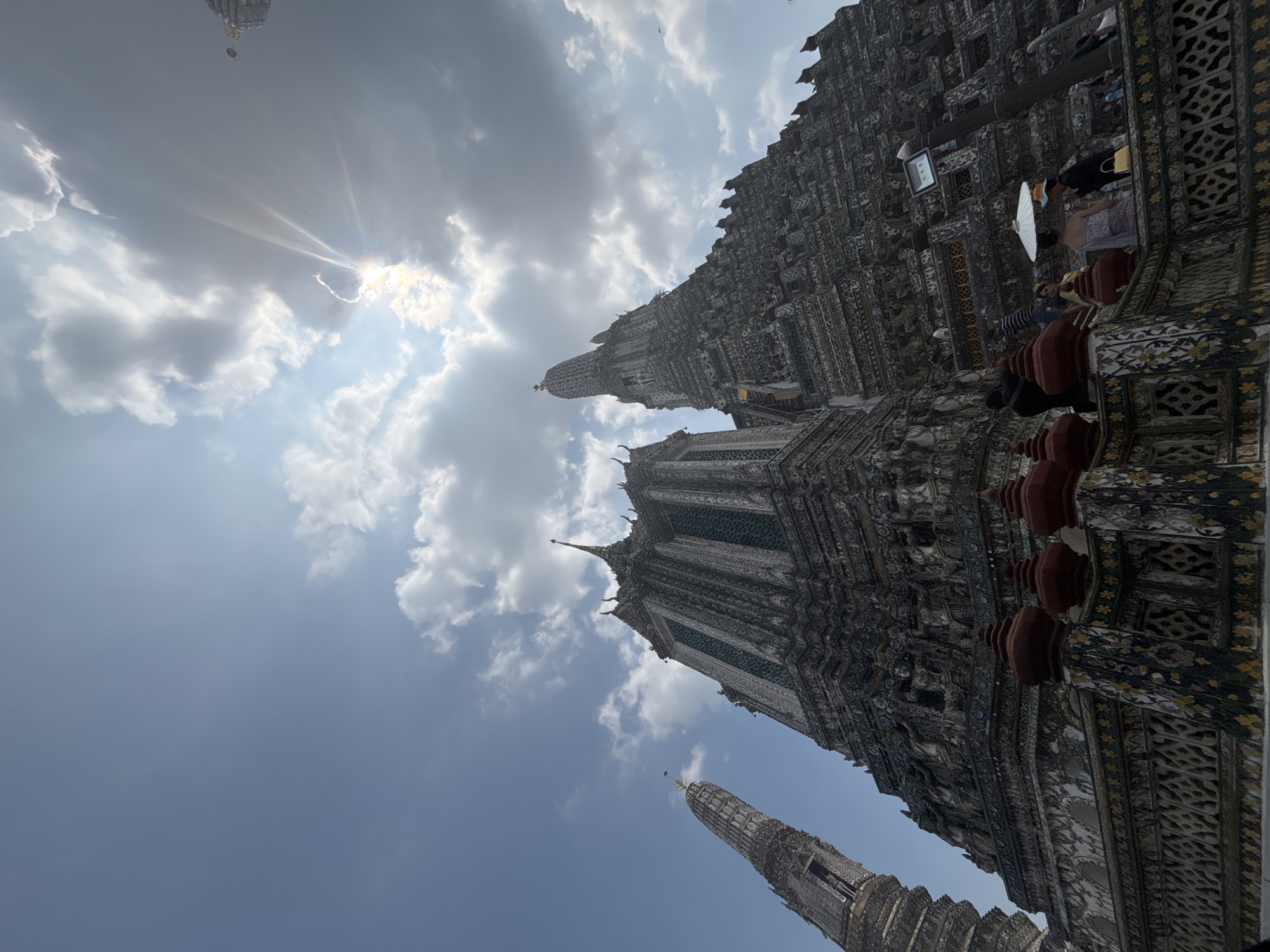 The image shows a low-angle view of Wat Arun Ratchawararam Ratchawaramahawihan, a Buddhist temple (wat) in Bangkok Yai district of Bangkok, Thailand. The temple is on the west bank of the Chao Phraya River. The photograph includes several spires of the temple against a sky filled with clouds, and partially obscured by the sun. Some people are visible on the temple structure.