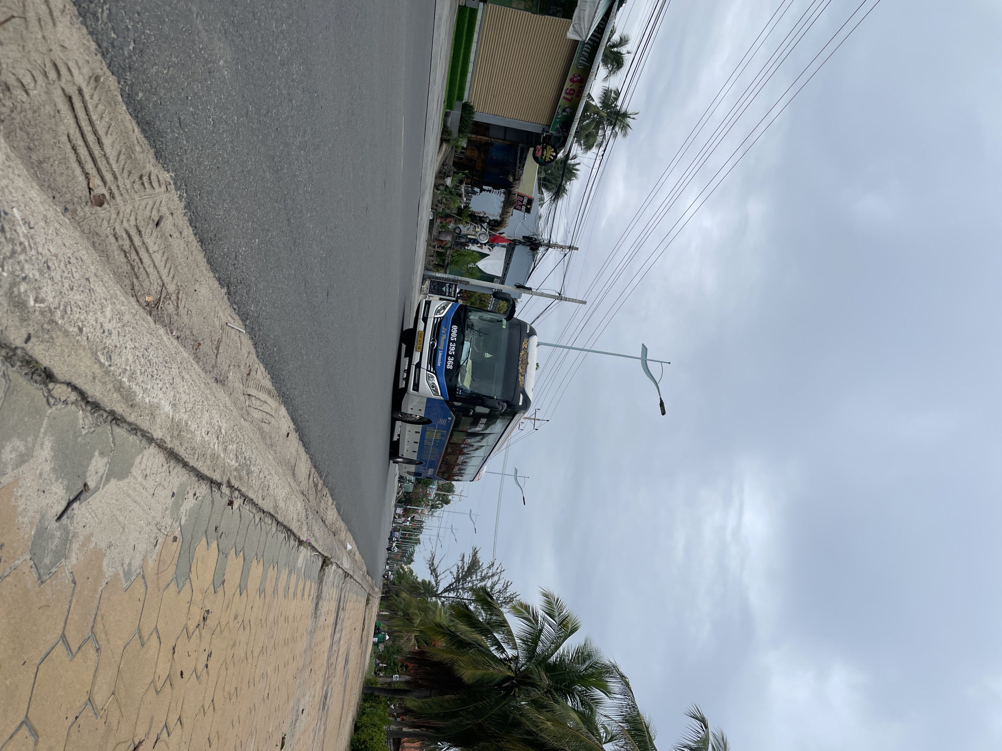The image shows a street scene in Phan Thiet Ward, Vietnam. A white and blue bus is driving on the road. The road has a sidewalk with hexagonal patterns. There are palm trees and buildings alongside the road. Power lines stretch across the sky, which is overcast.