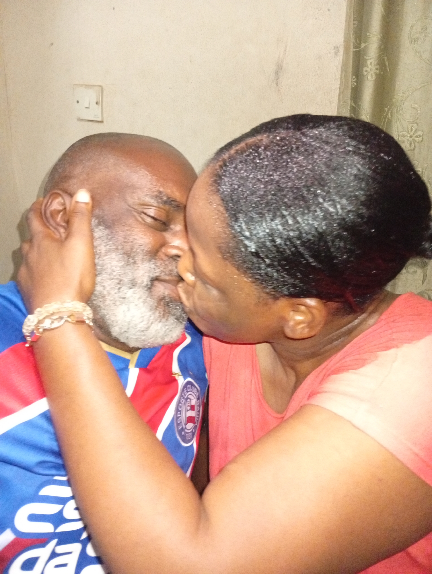 A woman kisses a man on the cheek. The man is dark-skinned with a bald head and a gray beard. He is wearing a blue and red sports jersey. The woman is dark-skinned with short, dark hair. She is wearing a pink shirt and a gold bracelet. She has her hand on the man's cheek. The background is a plain, light-colored wall. There is a light switch on the wall. To the right of the couple is a light-colored curtain. Based on the context provided, the location is Sabon Tasha, Nigeria.