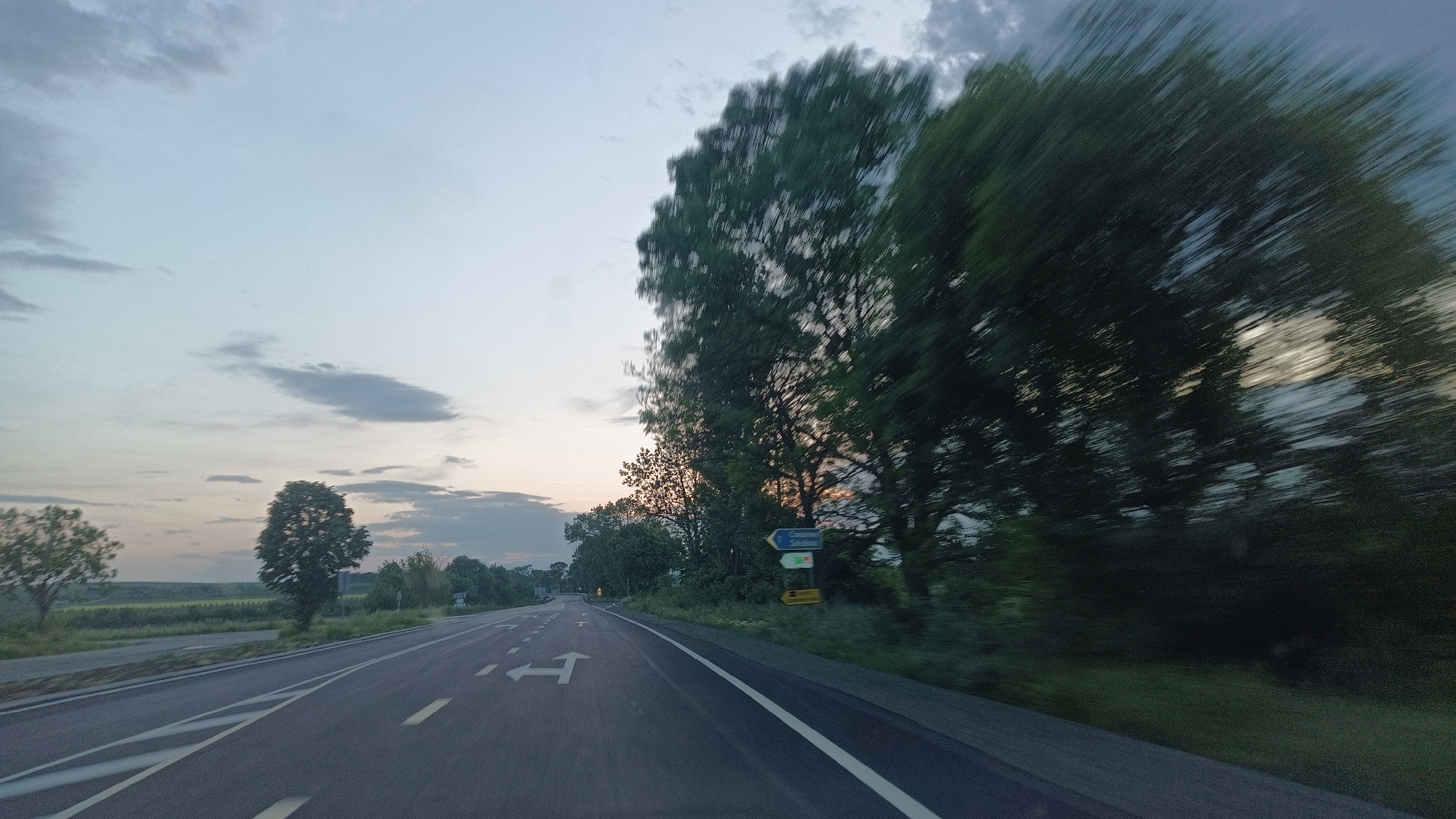 The image shows a two-lane road with white directional markings. The sky is overcast with some clouds. On the left side of the road is a grassy area with trees. On the right side, there are trees and a road sign indicating directions to "Sokolovo 1 km" and "Zlatintsa". The image appears to be taken from a moving vehicle, as some of the trees on the right side of the road are blurred. The overall scene suggests a rural or semi-rural environment, likely a road approaching or leaving the city of Sokolovo, Bulgaria.
