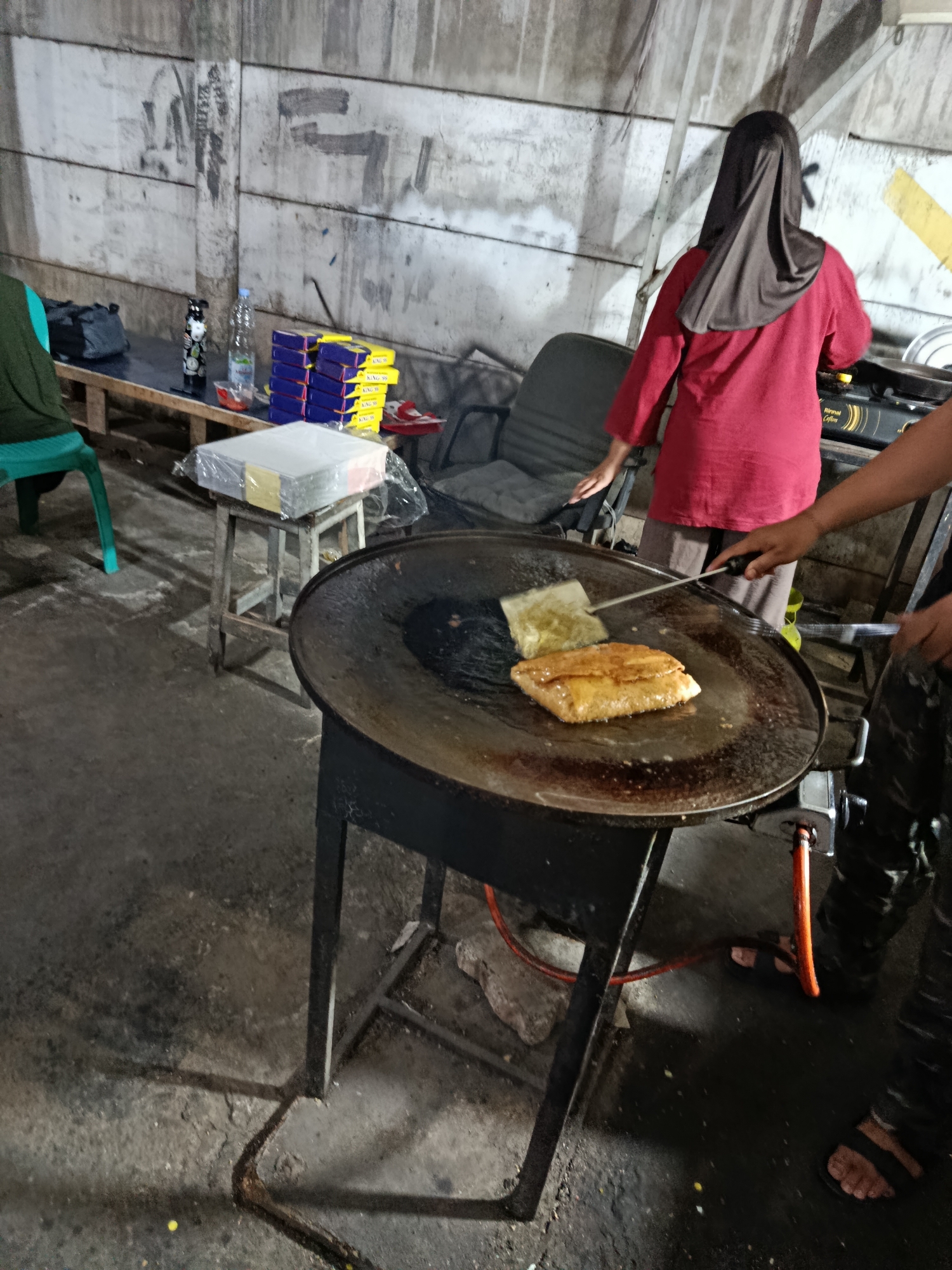 The image depicts an outdoor food stall in Depok, Indonesia. A woman wearing a red shirt and a dark hijab is cooking on a large, flat griddle. She is using a spatula to flip a golden-brown, folded pastry. The griddle is supported by a black metal stand, with a gas cylinder visible at the base. In the background, a concrete wall is visible, marked with what appears to be graffiti. Various items are arranged on a wooden table, including stacks of boxes, a water bottle, and a thermos. A plastic-wrapped stack of paper is placed on a small wooden stool. The ground is concrete, with some debris scattered around. The lighting suggests it is either evening or a dimly lit indoor/covered area. Another person's arm is visible on the right side of the frame, also involved in the cooking process.