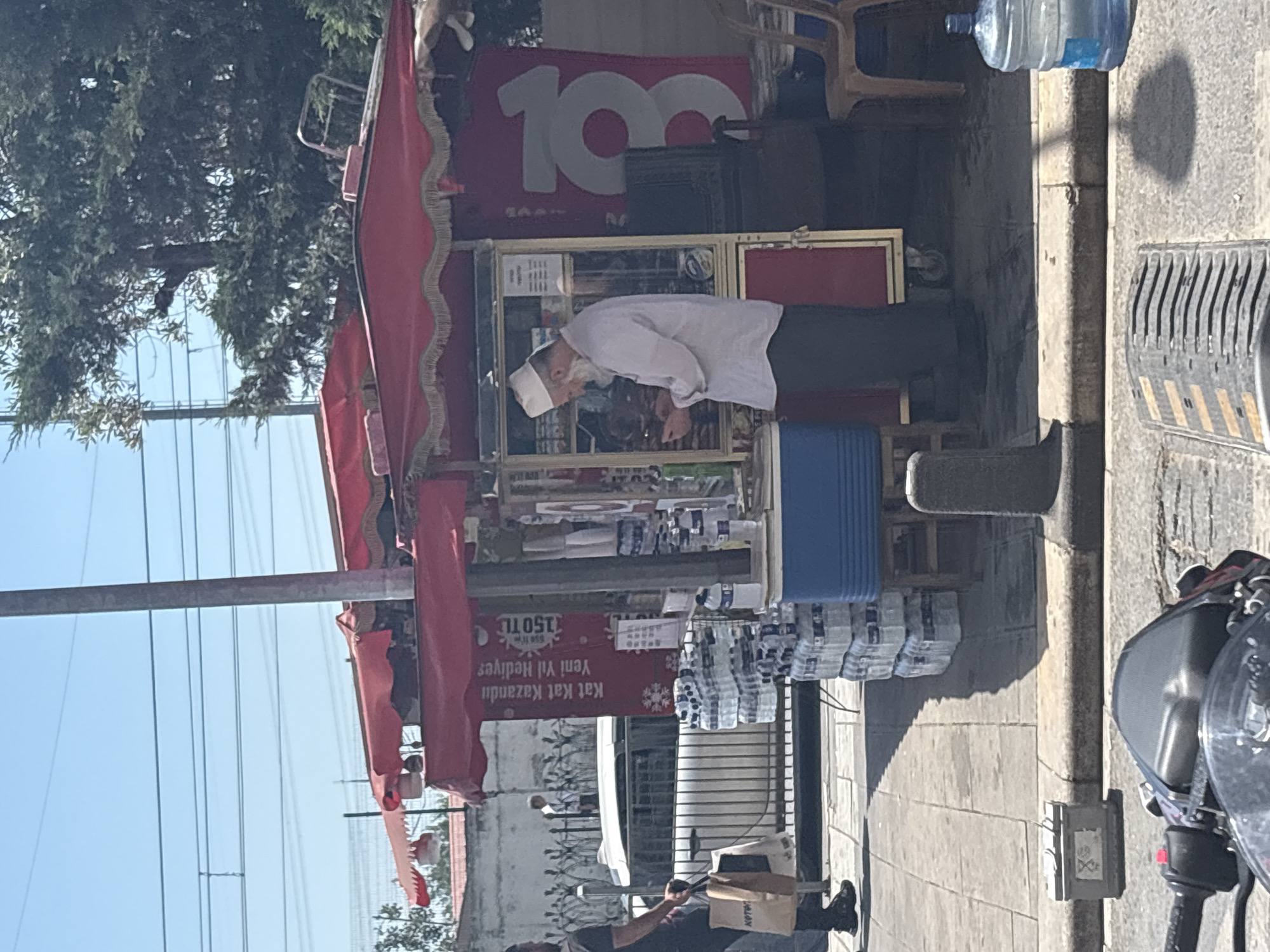 This image depicts a bustling outdoor street scene in an urban area of Gaziosmanpaşa, Turkey, under bright daylight with a clear blue sky. The central focus is a red street kiosk, covered by a large red canopy with gold-fringed trim. The kiosk features a glass display case on its left side and a solid red panel on its right. A red banner with white Turkish text is affixed to the front, reading "Kat Kat Kazandıran Yeni Yıl Hediyesi" (Winning Multiple New Year Gifts) and displaying "150 TL" along with a snowflake graphic. Another red banner on the right side of the kiosk shows a large white number "103". Standing behind the kiosk, on the left side, is an older man with a white beard and a white skullcap, wearing a light-colored long-sleeve shirt and dark pants. He is looking down at a small object held in his hands. To his left, on the sidewalk, is a blue insulated cooler, and stacks of bundled small plastic water bottles are arranged on the ground and on a metal stand. More large plastic water bottles are visible to the right of the kiosk. Two brown plastic chairs are situated on the sidewalk to the far right of the kiosk. On the left side of the image, a man wearing a dark short-sleeve shirt, dark shorts, and dark sandals is walking away from the kiosk towards the left, carrying two paper bags in his left hand, one visibly branded "KOTON". In the background, behind a decorative black metal fence, a white car is visible on the street. A distant, lightly dressed figure can be seen walking behind the car. Overhead, multiple power lines traverse the blue sky. A tall, gray utility pole stands between the two red banners of the kiosk. Green trees and foliage are visible in the background, particularly on the right side. The foreground shows parts of a dark-colored motorcycle or scooter, including the handlebars and front panel, indicating the perspective of the photograph. The sidewalk is paved with concrete slabs, and a gray concrete curb separates it from the asphalt street. A small, square, gray utility box rests on the sidewalk near the curb. In the immediate foreground, a gray metal object with horizontal ridges, possibly a speed bump or drainage cover, is on the road surface. Shadows from the kiosk and various objects are distinctly cast on the sunny