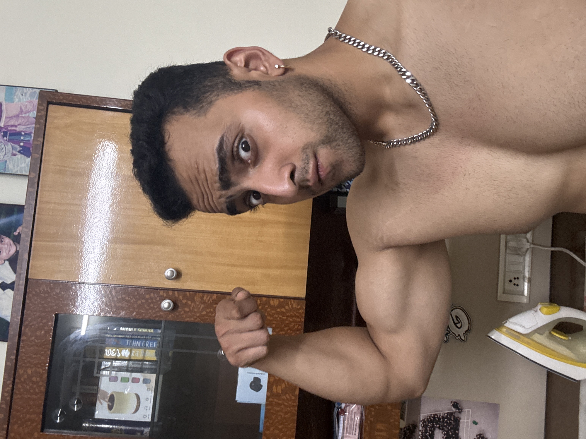 A shirtless man with a silver chain around his neck flexes his arm in what appears to be an indoor setting. A wardrobe or cabinet stands behind him, adorned with framed pictures and what appears to be a book collection visible through a glass front. Adjacent to the man is an iron, suggestive of a domestic environment. While the photo provides clues about the scene, there's no information to indicate the location is Kolkata, India. The specific city location cannot be determined from the image.