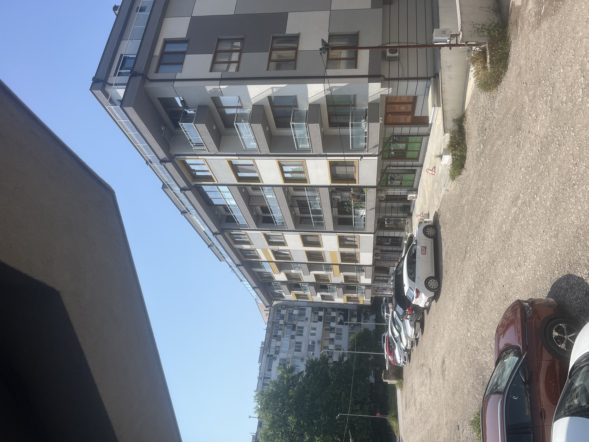 This image depicts a sunny daytime scene in Varna, Bulgaria, showcasing a contemporary urban environment. The primary subject is a multi-story modern residential building in the foreground, characterized by a distinctive facade with alternating grey and white panels. It features numerous balconies with clear glass railings and various windows, some accented with yellow frames. Several air conditioning units are visible on the building's exterior.
Adjacent to this building, an unpaved parking area extends into the foreground, where several cars are parked. A white compact car, possibly a hatchback, is prominent, with the word "EXIT" clearly visible on its rear window. Other cars, including a red-brown SUV, are parked nearby. A paved walkway runs alongside the modern building, where some low-lying green bushes are planted.
In the mid-ground, behind the modern building and partially obscured by trees, stands an older, taller apartment block. This building appears denser, with many windows and balconies, typical of residential structures in the region. The clear blue sky above suggests fair weather conditions. The scene is static, with no human activity observed, presenting a common street view of residential buildings. The overall impression is one of a quiet, everyday urban setting.