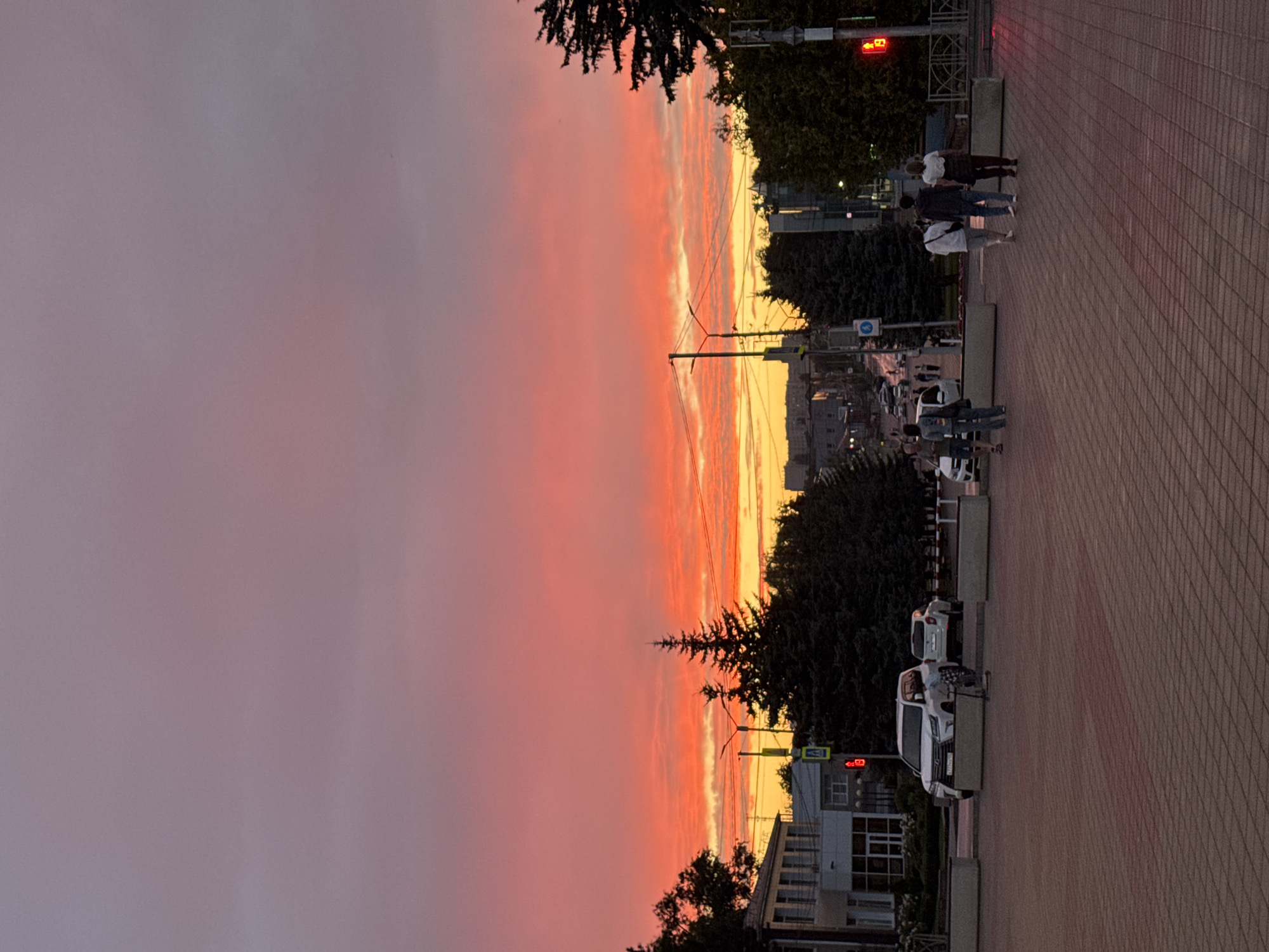 The image depicts a sunset over the city of Mikhaylovsk, Russia. The sky is filled with vibrant hues of orange, pink, and purple, indicating a dramatic end to the day. In the foreground, a paved walkway with a red brick pattern leads into the scene. Several people are walking on the walkway, some in groups and some individually. To the left, a white SUV is parked, and further down the street, more cars are visible, suggesting a typical urban environment. The street is lined with trees, including several tall, dark evergreen trees. Buildings of various sizes and styles are scattered throughout the background, some appearing to be residential and others commercial. Traffic lights and street signs are also present, indicating the presence of regulated city streets. The overall atmosphere of the image is serene and picturesque, capturing the beauty of a sunset in a Russian city.