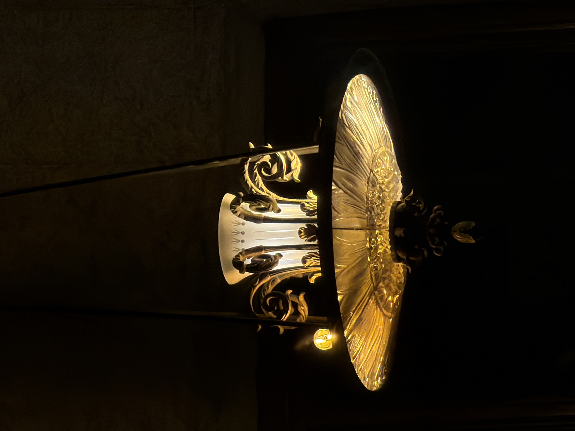 The image depicts a decorative wall sconce in Saint Petersburg, Russia. The sconce features a central translucent glass column, adorned with ornate gold-colored metalwork in floral and scroll patterns. A circular, fan-like gold reflector projects outward, directing light. The immediate environment is dark, suggesting the photograph was taken in a dimly lit interior. The background texture indicates a wall surface.