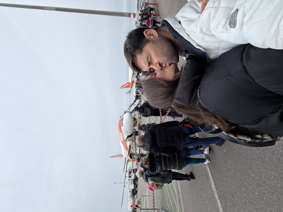 The image depicts an airport tarmac in Colombier-Saugnieu, France, likely Lyon-Saint-Exupéry Airport (LYS), under an overcast sky. In the immediate foreground, two individuals, a female with long brown hair wearing a black puffer jacket and a male with short brown hair and stubble wearing a white puffer jacket, are positioned right of center. They are engaged in a kiss, with the male's eyes visibly squeezed shut and his lips pursed.
Behind the couple, a queue of approximately 15-20 passengers is visible, standing on an asphalt tarmac marked with white lines and delineated by red and white striped barrier tape on the left. In the midground, two orange and white EasyJet aircraft are parked. The prominent aircraft, an Airbus A320 type, is being boarded by passengers via a mobile airstair at its front left door. A ground service vehicle and a ground crew member in an orange high-visibility vest are positioned near this aircraft's nose. A second EasyJet aircraft is partially visible further back on the right. A tall, slender grey pole stands vertically on the far right side of the frame.