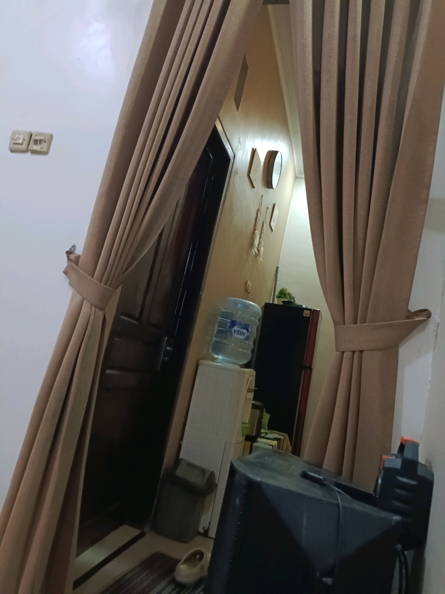 The image depicts an indoor scene, likely a residential room, in Rancang Kencono, Indonesia. The focal point is a set of beige curtains that are pulled to the sides, revealing an interior doorway and a section of a room. On the wall to the left of the curtains, there are two white electrical outlets. The room beyond the curtains appears to contain a white cooler with a large water dispenser bottle on top, a dark-colored refrigerator, a small potted plant, and a large black speaker or audio equipment in the foreground. The floor visible near the doorway shows a dark rug and a light-colored shoe. The overall lighting suggests it is daytime, with artificial light sources also potentially active. There are no people visible in the image.