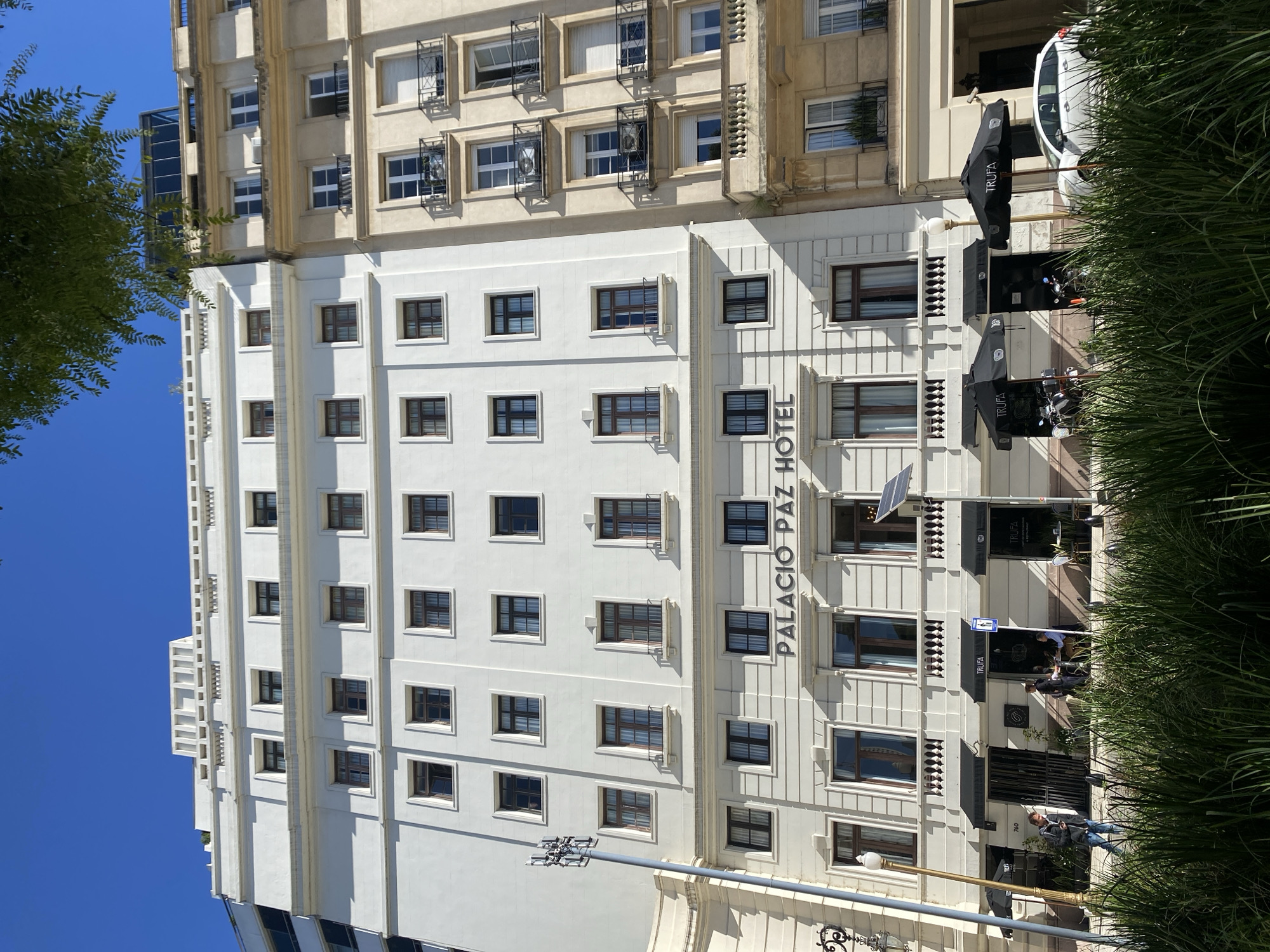 This media file captures the exterior of the Palacio Paz Hotel in Buenos Aires, Argentina, under a bright, clear blue sky. The primary subject is the multi-story hotel building, which exhibits two distinct architectural styles.
The lower and mid-section of the building features a light, possibly white or cream-colored facade, characterized by symmetrical rows of dark-framed rectangular windows and decorative vertical fluting. The name "PALACIO PAZ HOTEL" is displayed prominently in dark, vertical lettering on this section. Below this, the word "HOTEL" is also partially visible horizontally.
Above the lighter section, an older, beige or tan-colored upper portion of the building extends, presenting a different style with more ornate window designs. Many windows on this upper section have visible air conditioning units, often protected by black metal grates or cages.
At street level, the ground floor of the hotel hosts outdoor commercial spaces. Two large, black umbrellas, each marked with the text "TRUFA" in white, define an outdoor seating area, likely for a café or restaurant. Several people are visible: some are seated at tables under the umbrellas, while others are walking past or standing near the hotel entrance. A white van or car is partially seen behind one umbrella, and a motorcycle is parked nearby.
Lush green foliage, including a tall leafy tree on the left and a row of spiky green plants along the bottom right, partially frames the building and the street scene. The strong sunlight and clear shadows indicate it is daytime. Street furniture, such as a lamppost equipped with a small solar panel and a street sign, further define the urban setting of Buenos Aires.