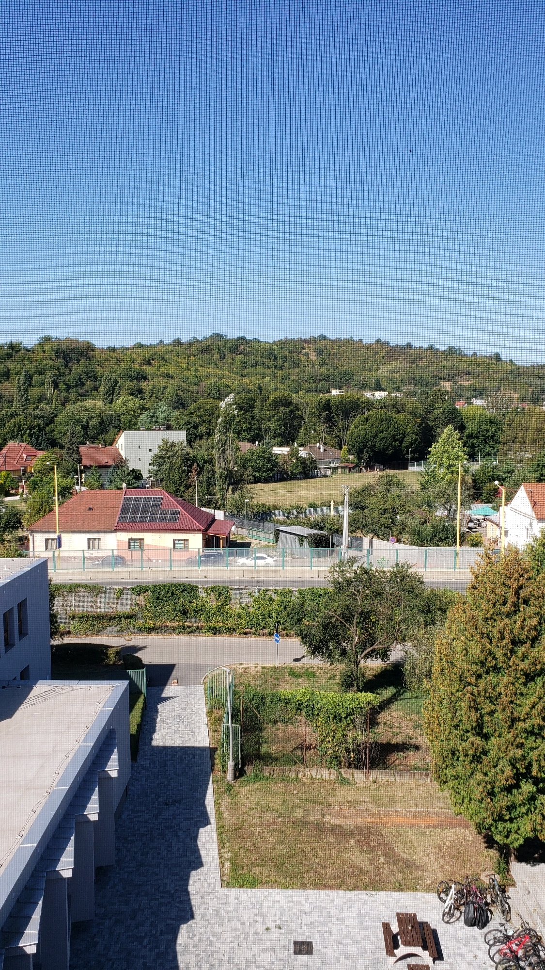 The image shows an elevated view of a residential area with a wooded hillside in the background. The foreground features the corner of a modern building with a flat roof and grey cladding. To the right, a paved walkway leads to a grassy area with a picnic table and several bicycles parked nearby. A road with a green fence runs horizontally across the mid-ground, separating the buildings from the forested landscape. Several houses with red tile roofs are visible, one of which has solar panels installed. The sky is clear and blue. The scene appears to be a typical suburban or semi-rural environment in Prešov, Slovakia.