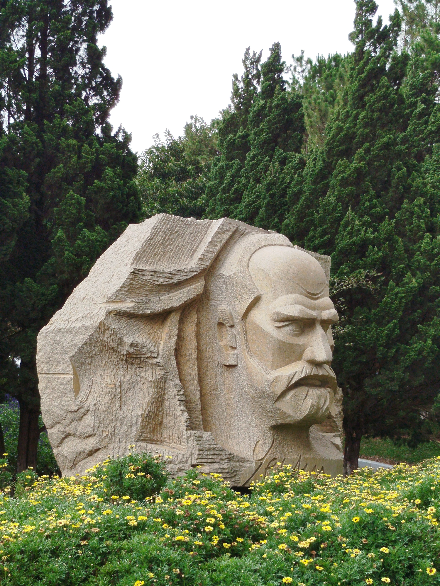 A large, monumental sculpture of a stern-faced man is the focal point of the image. The sculpture is carved from a light-colored stone. The backdrop features dense evergreen trees, suggesting a park-like setting. In the foreground is a vibrant field of yellow daisies, which contrast with the stone of the sculpture and the green trees. This sculpture is located in the city of Da Lat, Vietnam.