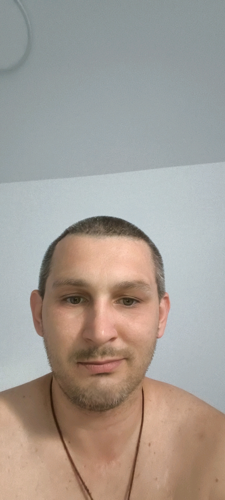 A man with short brown hair and a light beard is pictured from the chest up, facing the camera. He has light brown eyes and is looking directly at the viewer. A thin brown cord necklace is visible around his neck. He appears to be indoors, with a plain, light-colored wall and ceiling in the background. There are no other individuals or objects of note in the frame.