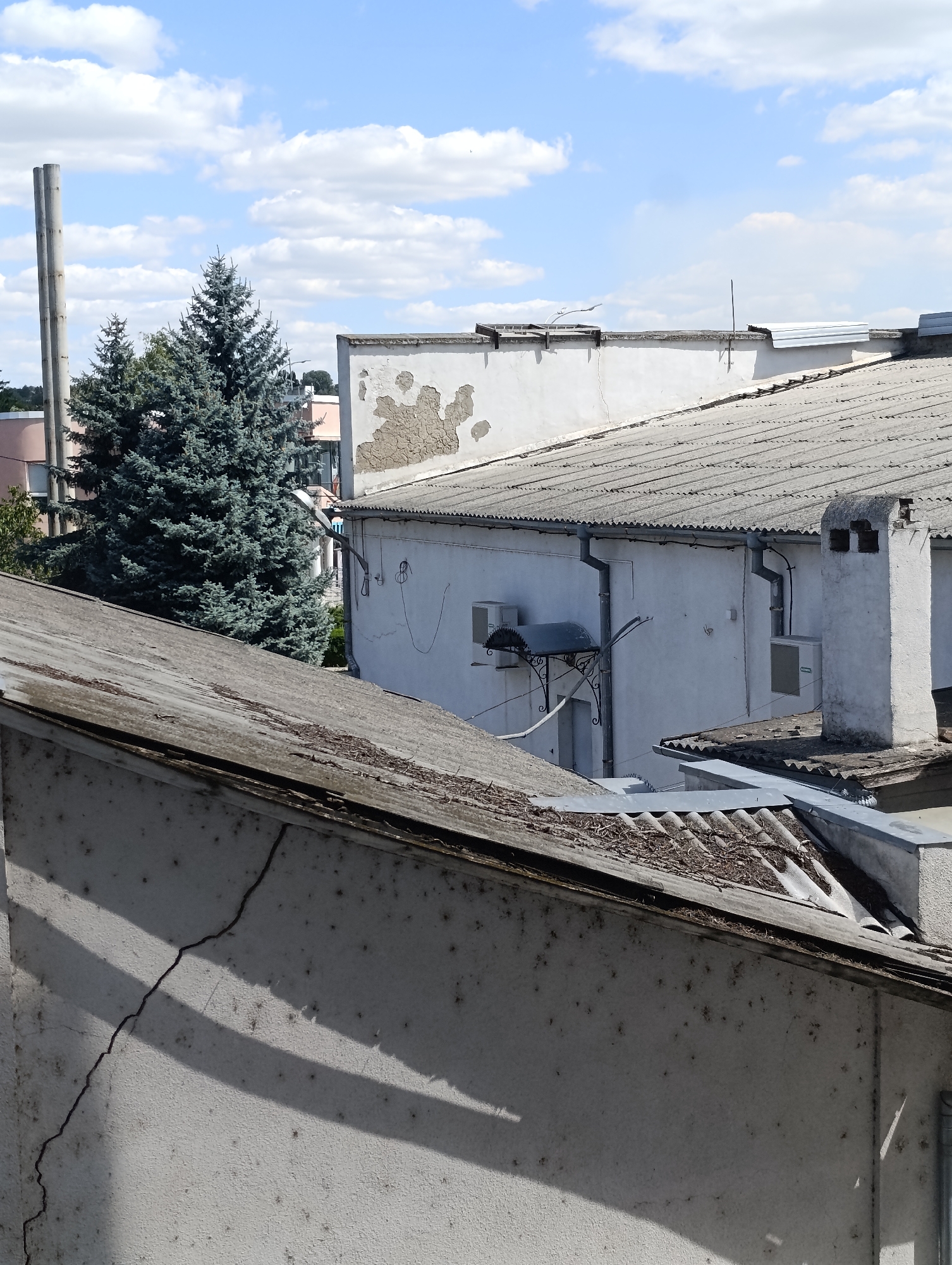 The image captures a view of rooftops and surrounding elements under a partially cloudy blue sky in Dubăsari town council, Moldova. In the foreground, a weathered, grey corrugated roof is visible, with visible signs of decay and debris. Adjacent to this is a white wall with a significant crack running vertically, marked by shadows from an unseen source. This wall is also speckled with numerous dark marks. Beyond this, a white building with a grey, corrugated roof is seen, featuring a visible chimney with two openings and air conditioning units mounted on its exterior. Further in the background, a dense cluster of blue-green fir trees stands tall, contrasting with the predominantly grey and white urban structures. A tall, grey cylindrical structure, possibly a smokestack or pipe, rises behind the trees. The overall scene depicts an industrial or commercial area, showing signs of age and weathering. There are no people or animals visible in the image.