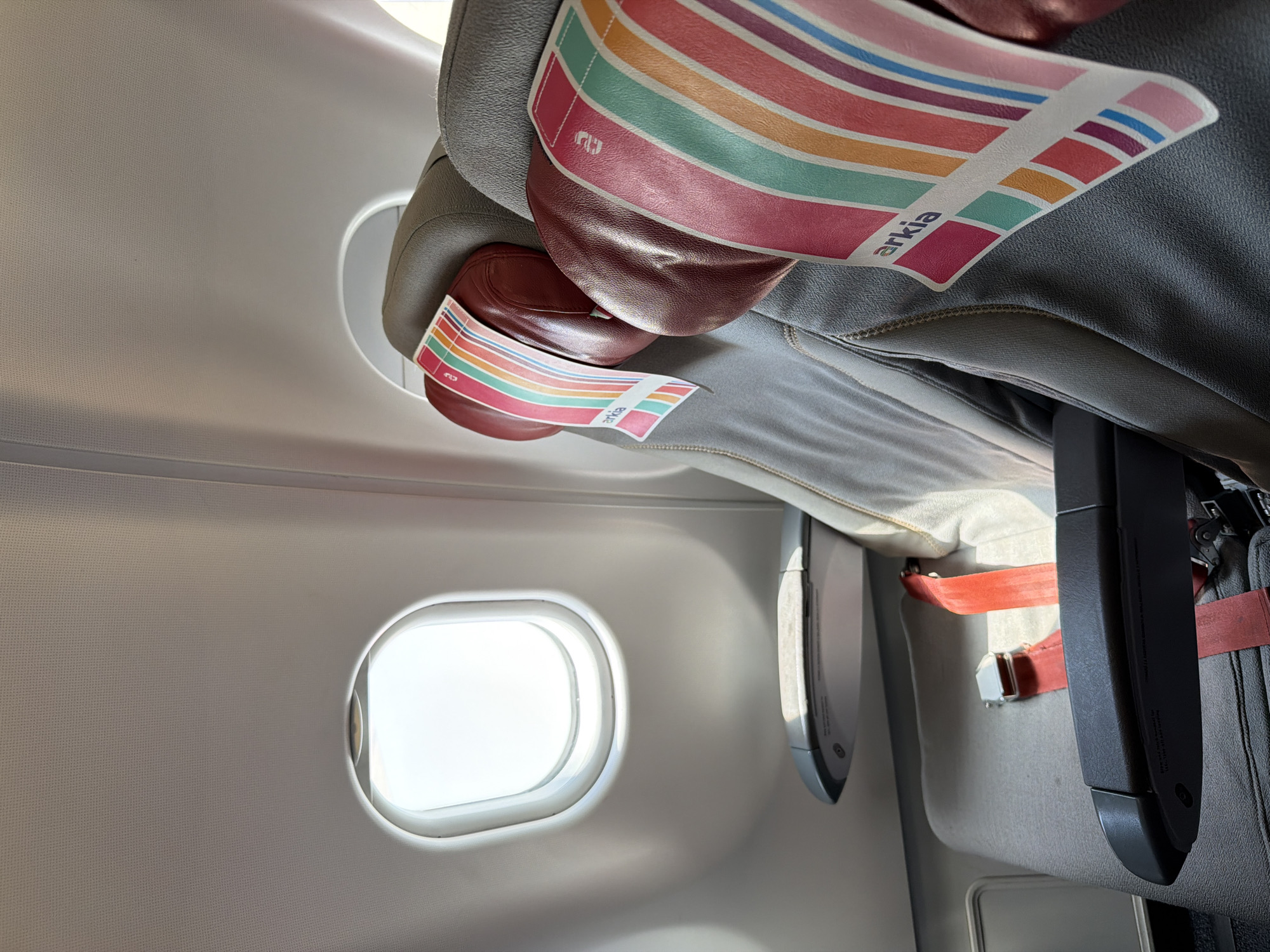 The image shows the interior of an airplane cabin. A window is visible on the left side of the frame, allowing bright light to enter. An airplane seat is present, with an "arkia" branded headrest cover. A seatbelt and a portion of a suitcase are also visible. There is no indication of the image's location being in Tzafria, Israel.