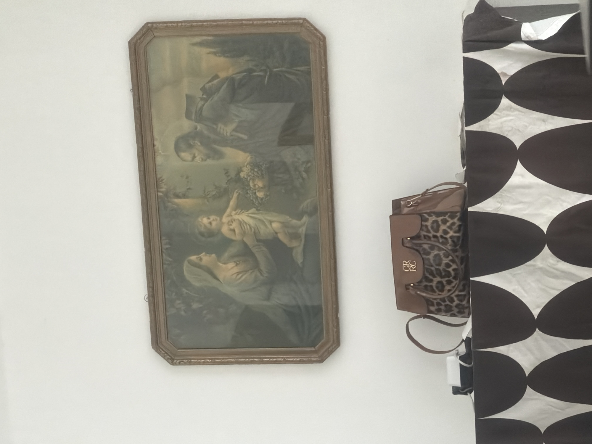 The image shows a framed painting of a religious scene, possibly the Holy Family, hanging on a white wall. In the foreground, a patterned tablecloth is draped over a surface. On top of the tablecloth are a handbag with leopard print accents and a small white object. The painting depicts a woman in a blue robe holding a child, with a man in a brown robe standing to the right. There is a basket of fruit near the child. The overall scene is indoors, with artificial lighting. There is no specific information about the location in Mannheim, Germany, that can be deduced from the image.