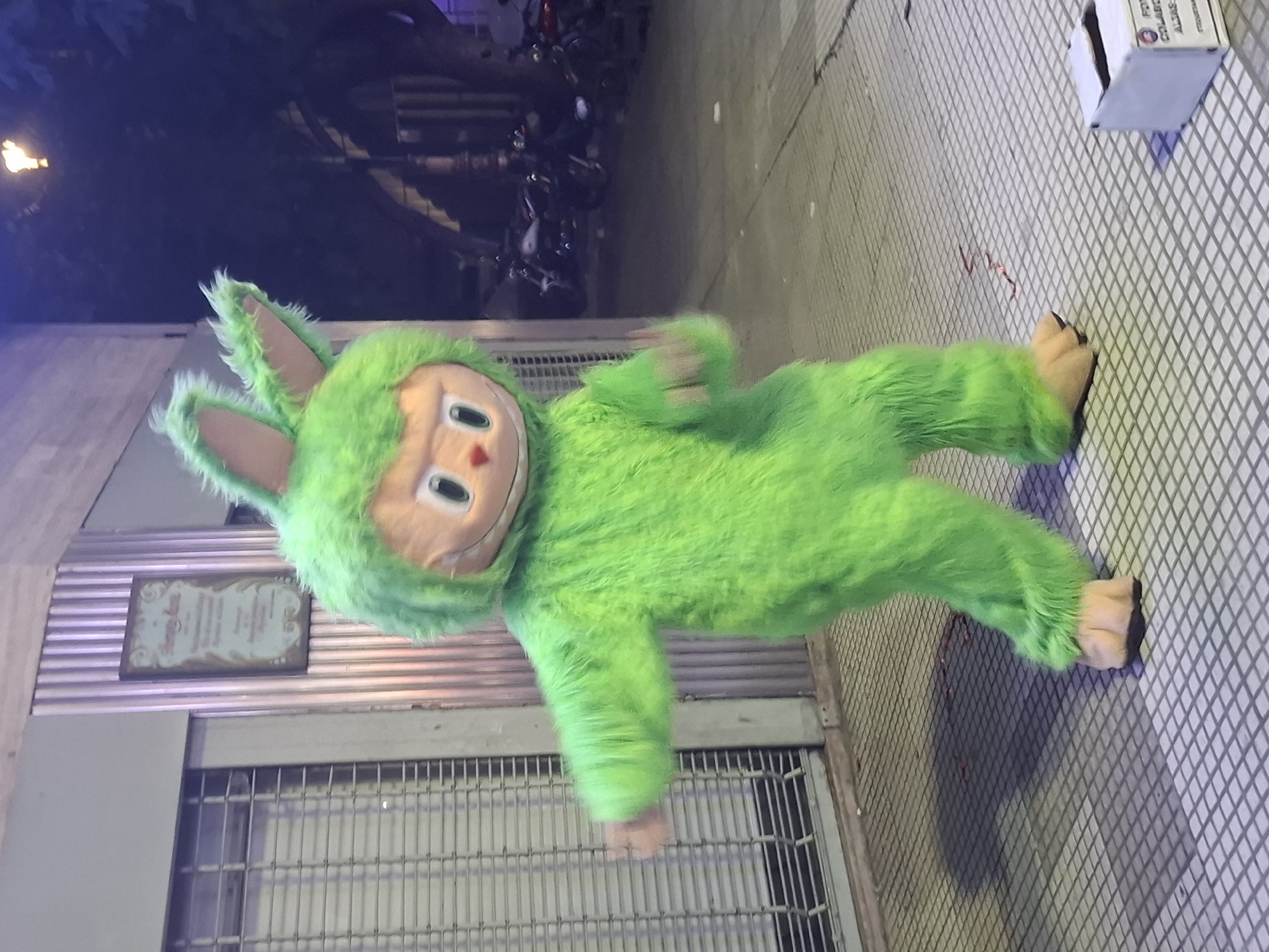 A green mascot costume resembling a furry creature with a cartoonish face lies on a sidewalk in Buenos Aires, Argentina. The mascot has long ears, a fuzzy green body, and large, light brown feet. It appears to be lying near a metal grate in front of a building with a sign. The sidewalk is tiled with a small white box sitting on it near the top right corner. In the background, blurred images of parked motorcycles are visible, as well as a street light.