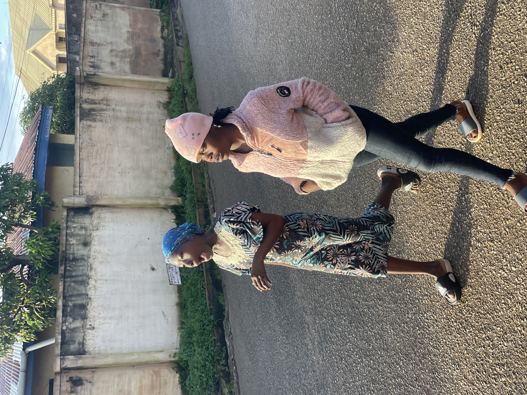 Two young women are walking on a paved road in Jos, Nigeria. The woman on the left is wearing a blue headscarf and a floral print dress, with black slides on her feet. She appears to be mid-conversation or exclaiming something, with her mouth open. The woman on the right is wearing a pink and white fuzzy jacket, dark jeans, and grey slides. She has a pink beanie on her head with some drawn-on designs. They are walking away from the camera towards the right side of the frame. Behind them is a textured, light-colored wall with some dark staining, and beyond that, vegetation and buildings with corrugated metal roofs. The lighting suggests it is late afternoon, with long shadows cast on the road.
