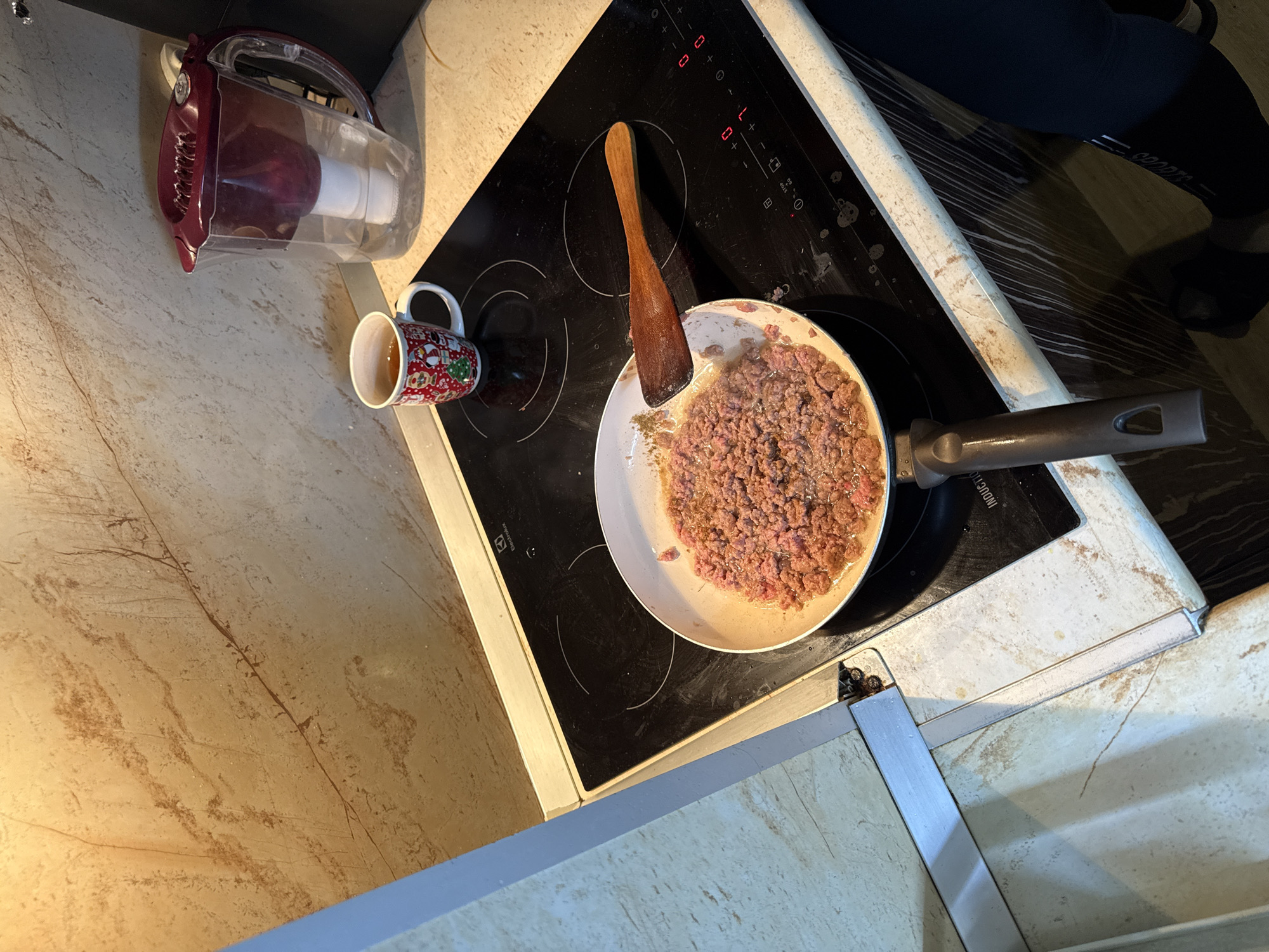 The image shows a close-up view of a kitchen scene. On a black induction stovetop, a white frying pan is filled with raw ground meat that appears to be cooking. A wooden spatula rests on top of the frying pan. To the right of the pan, a white mug decorated with Christmas-themed figures is filled with a dark liquid, likely tea or coffee. Next to the mug is a red and clear water filter pitcher, with some water visible inside. The stovetop has illuminated control buttons. The backsplash behind the stovetop is made of light-colored, marble-patterned tiles. The countertop is also a light-colored material with a marble pattern. The overall impression is a domestic setting where cooking is in progress. The location is not specified within the image itself, and therefore cannot be determined to be in Stara Zagora, Bulgaria.