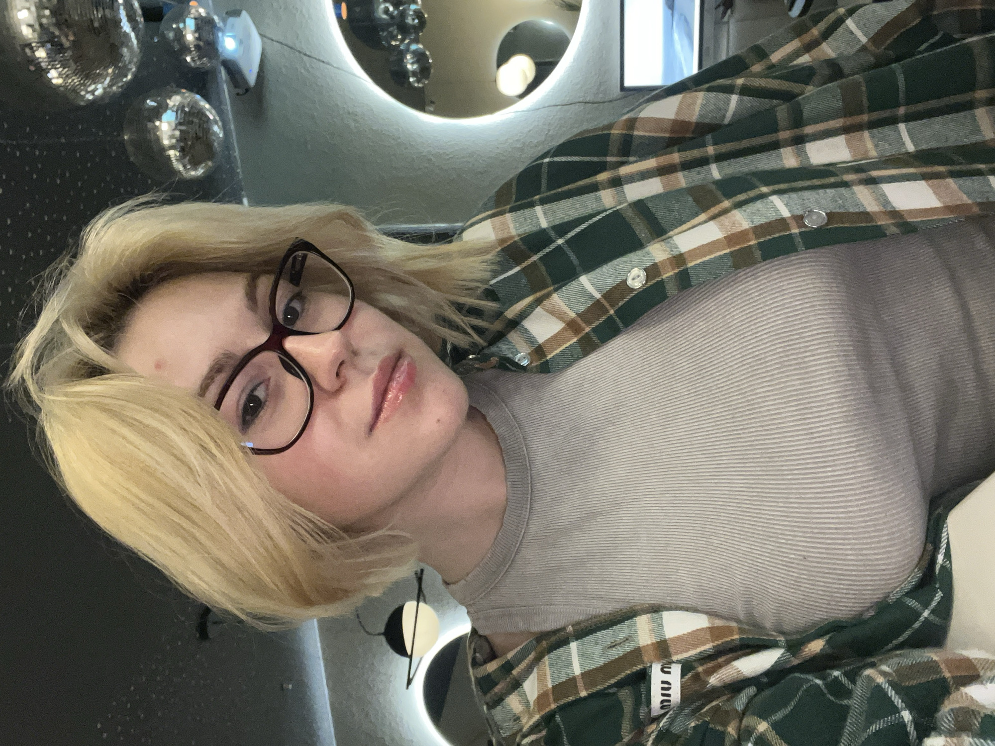 A woman with blonde hair and glasses is posing for a selfie in an indoor setting. She is wearing a grey ribbed top and a green, brown, and white plaid shirt. There are disco balls in the background on the left. A circular light is also in the background. The brand label on the shirt near the bottom reads "Miu Miu." The location is suggested to be Rafailovići, Montenegro.