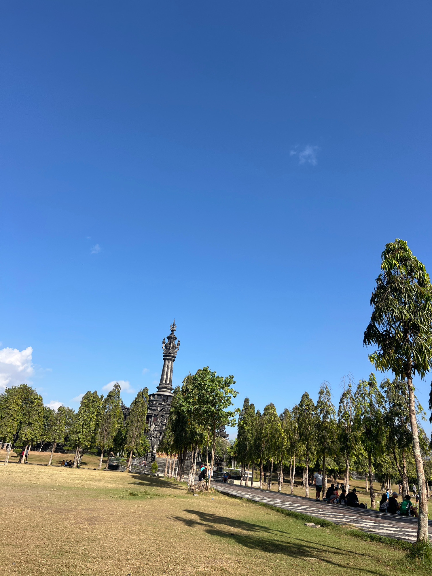 The image depicts a monument and a row of trees under a clear blue sky. The monument, which appears to be made of dark stone, has an intricate design with several levels and a pointed top. It is situated in the background, behind a line of evenly spaced, tall, slender trees with lush green foliage. A pathway runs in front of these trees, and several people are seen sitting or standing along it.
In the foreground, a large grassy area with dry, brown grass is visible, casting shadows from the trees. The overall environment suggests an outdoor public space, possibly a park or a monument ground. The scene is set in Denpasar, Indonesia, a location known for its cultural landmarks and lush greenery. The presence of people suggests it is a place visited by locals or tourists. The lighting indicates it is daytime, with the sun high in the sky, casting distinct shadows.