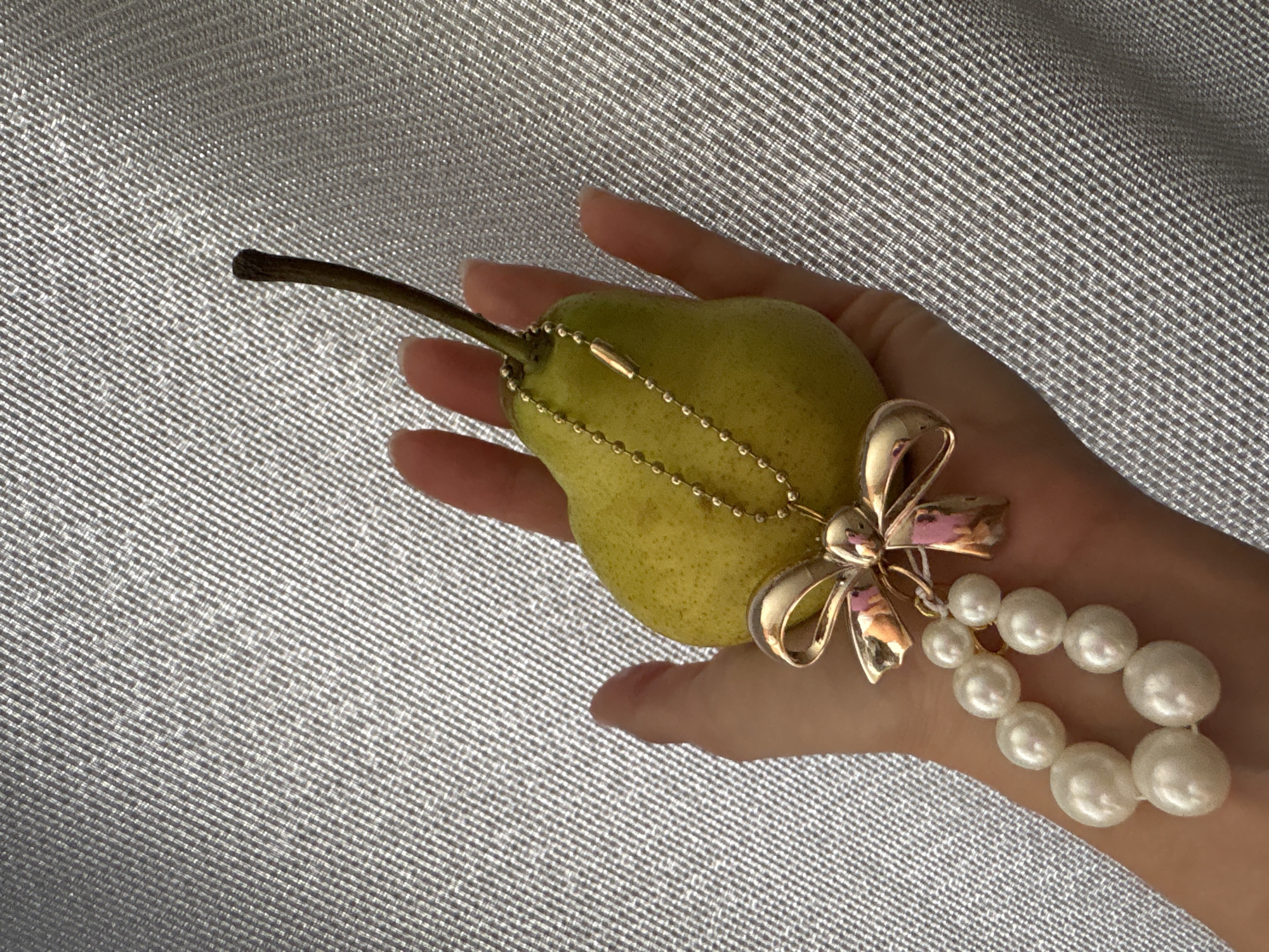 A hand holds a green pear, adorned with a gold chain, a gold bow, and a pearl bracelet. The background is a textured silver fabric. The location is unknown, and there is no evidence to suggest it is in Chaikovsky, Russia.