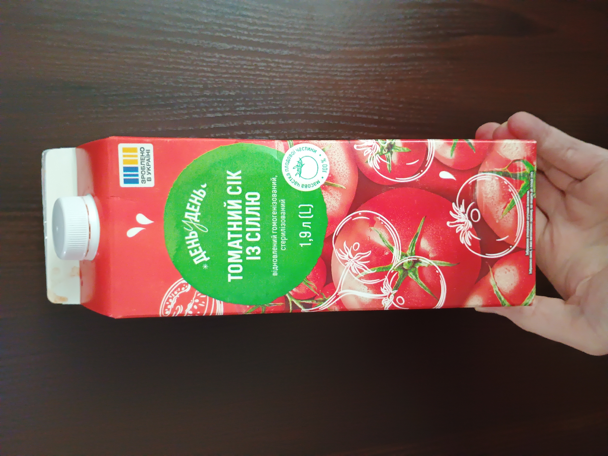 The image shows a hand holding a red carton of tomato juice. The carton has a white screw-top lid on the left side. The carton is mostly red with a large green circular label on it. Inside the green circle, there is text in Ukrainian, reading "ДЕНЬ ДЕНЬ." and "* ТОМАТНИЙ СІК ІЗ СІЛЛЮ". Below this, it says "відновлений гомогенізований, стерилізований" and "1,9 л (L)". To the left of the green circle, there is a smaller yellow and blue logo with "ЗРОБЛЕНО В УКРАЇНІ" written on it. The background of the carton features illustrations of tomatoes. There is also a circular stamp with a drawing of a sprout and the text "частка плодової частини" and "100%". The image appears to be taken indoors, on a wooden surface. The lighting suggests it is daytime. There is no specific weather information, but the overall scene is casual and everyday. The hand is holding the carton horizontally, suggesting it might be presenting the product. The visible text indicates the product is Ukrainian-made tomato juice with salt, reconstituted, homogenized, and sterilized, with a volume of 1.9 liters.
