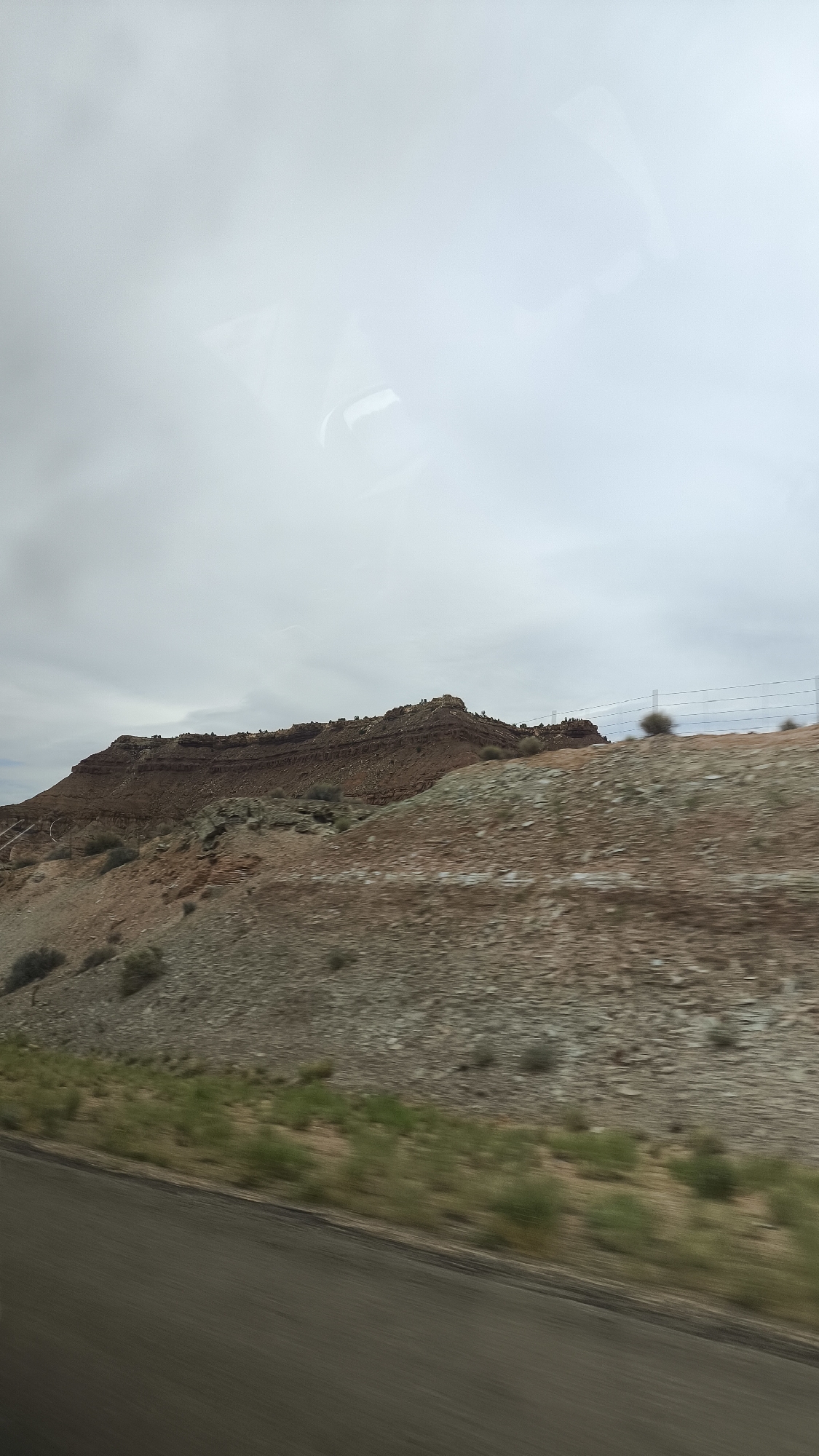 The image shows a panoramic view of a desert landscape with red rock formations under a cloudy sky. The foreground features a blurred road and a grassy embankment. The mid-ground consists of rolling hills covered in rocky terrain and sparse vegetation. In the background, a large mesa with distinct horizontal strata dominates the horizon. A wire fence is visible along the top of the mid-ground hills. The scene suggests a dry, arid environment, likely in a natural park or undeveloped area. The overall impression is one of vastness and geological history.