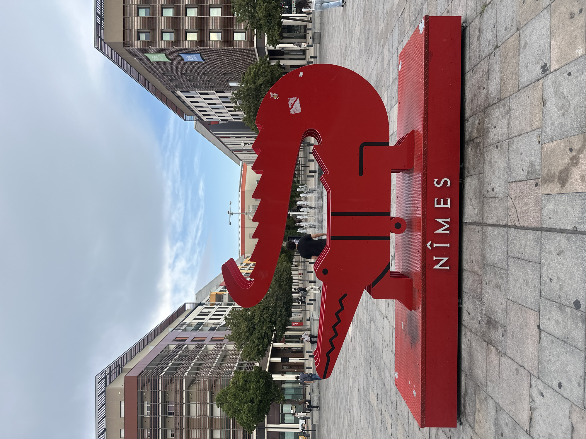 This is a vertical, medium-shot photograph of a bright red metal sculpture that resembles a stylized alligator or crocodile head. The sculpture is situated on a grey paved square, likely a public space, with several buildings in the background. The sculpture itself is vibrant and eye-catching, with a prominent snout, sharp teeth, and a rounded head. The letters "NÎMES" are emblazoned in white on the side of the sculpture's base, indicating the location.
In the background, modern buildings with repetitive window patterns and vertical wooden slats line the square. Some of these buildings have banners, including one that appears to say "ibis." Beyond the immediate area of the sculpture, there are trees and people walking in the distance. A fountain is also visible, with water jets creating a spray in the mid-ground. The people in the image are mostly casually dressed, and their activities appear to be leisurely, such as walking or standing around.
The sky is overcast with some wispy clouds, suggesting a daytime setting, possibly late afternoon or early evening given the softness of the light. The overall atmosphere is calm and urban. The sculpture appears to be a prominent landmark or piece of public art in the city of Nîmes, France. The focus of the image is the red sculpture, with the urban environment serving as a backdrop.