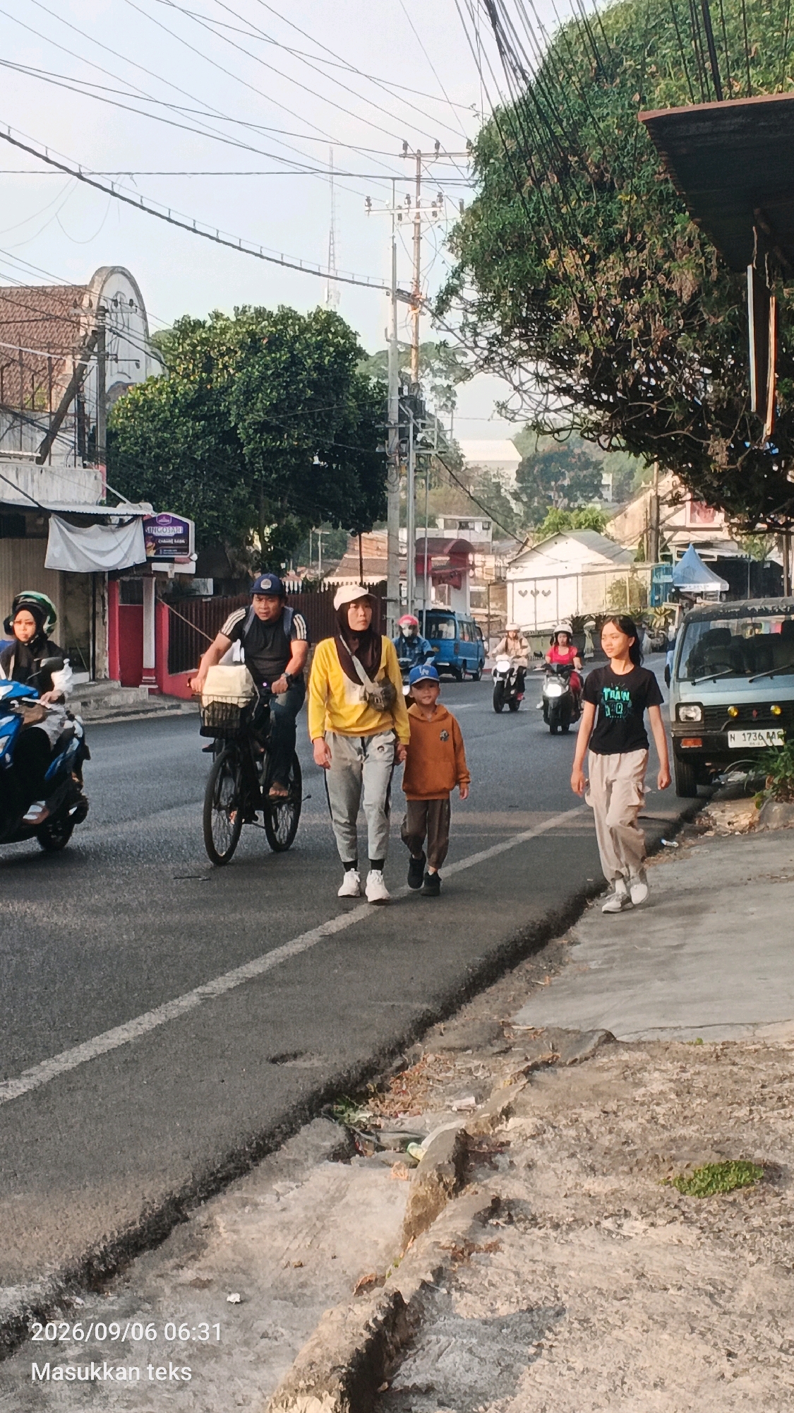 On a paved road in Malang, Indonesia, an early morning scene unfolds. A family is out for a walk. A man on a bicycle leads the way, with a basket of goods on the front. Behind him, a woman and a young boy walk hand-in-hand. The woman wears a yellow long-sleeved shirt and grey pants, while the boy is dressed in an orange hoodie and brown pants. To their right, a young woman in a black t-shirt and beige pants walks along the sidewalk, looking towards the right. In the background, several motorbikes are visible, one with a rider wearing a pink helmet and another with a rider in a blue helmet. A light blue van is parked on the right side of the road, and a light blue car is also present. The environment suggests a residential area with buildings and trees lining the street, under a bright, clear sky. The timestamp on the image indicates the date and time as 2026/09/06 06:31.