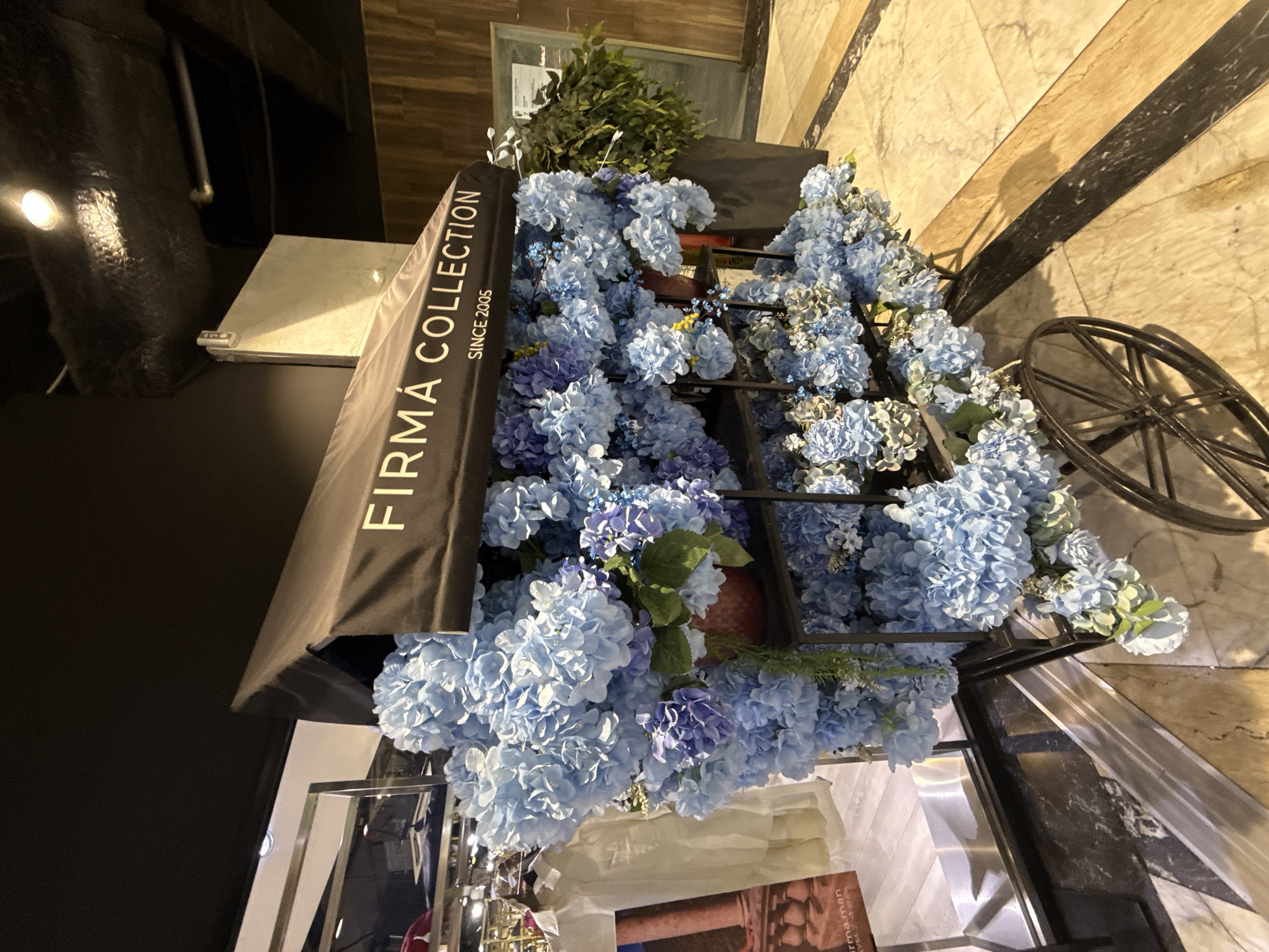 A decorative cart filled with light blue and purple hydrangeas is displayed in front of a store entrance. The cart has a black canopy with "FIRMA COLLECTION SINCE 2005" printed in white. The cart is positioned on a marble floor with black and tan tile accents. In the background, a large potted plant with dark green foliage is visible. To the left of the cart, a display mannequin wearing a light-colored garment is partially visible. The lighting suggests an indoor environment, possibly a shopping mall or boutique. The image does not provide any specific information about the city of Bishkek, Kyrgyzstan.