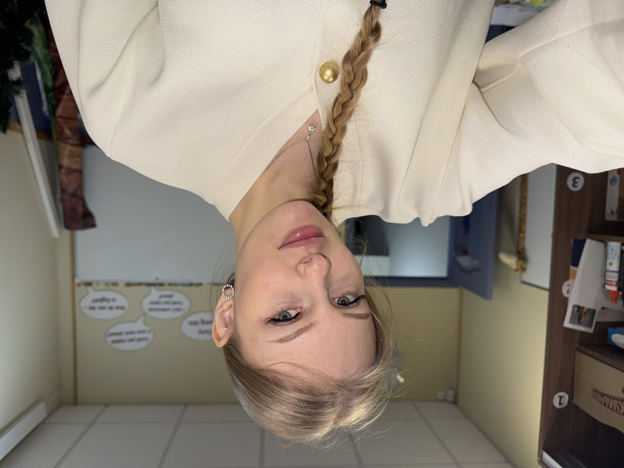 The image is a rotated selfie of a young woman with light blonde hair braided to the side. She is wearing a cream-colored cardigan with a gold button and a silver necklace. She has light eyes and pink lips. Her earring is a small, round, silver hoop.
The background appears to be an indoor setting, potentially a classroom or office. There is a whiteboard with speech bubble cutouts containing writing. There's also a wooden cabinet or shelving unit with numbered labels (1 and 3 visible) and some items on the shelves. The floor is tiled with light-colored, square tiles. The location in the city of Perm, Russia cannot be confirmed from the image.