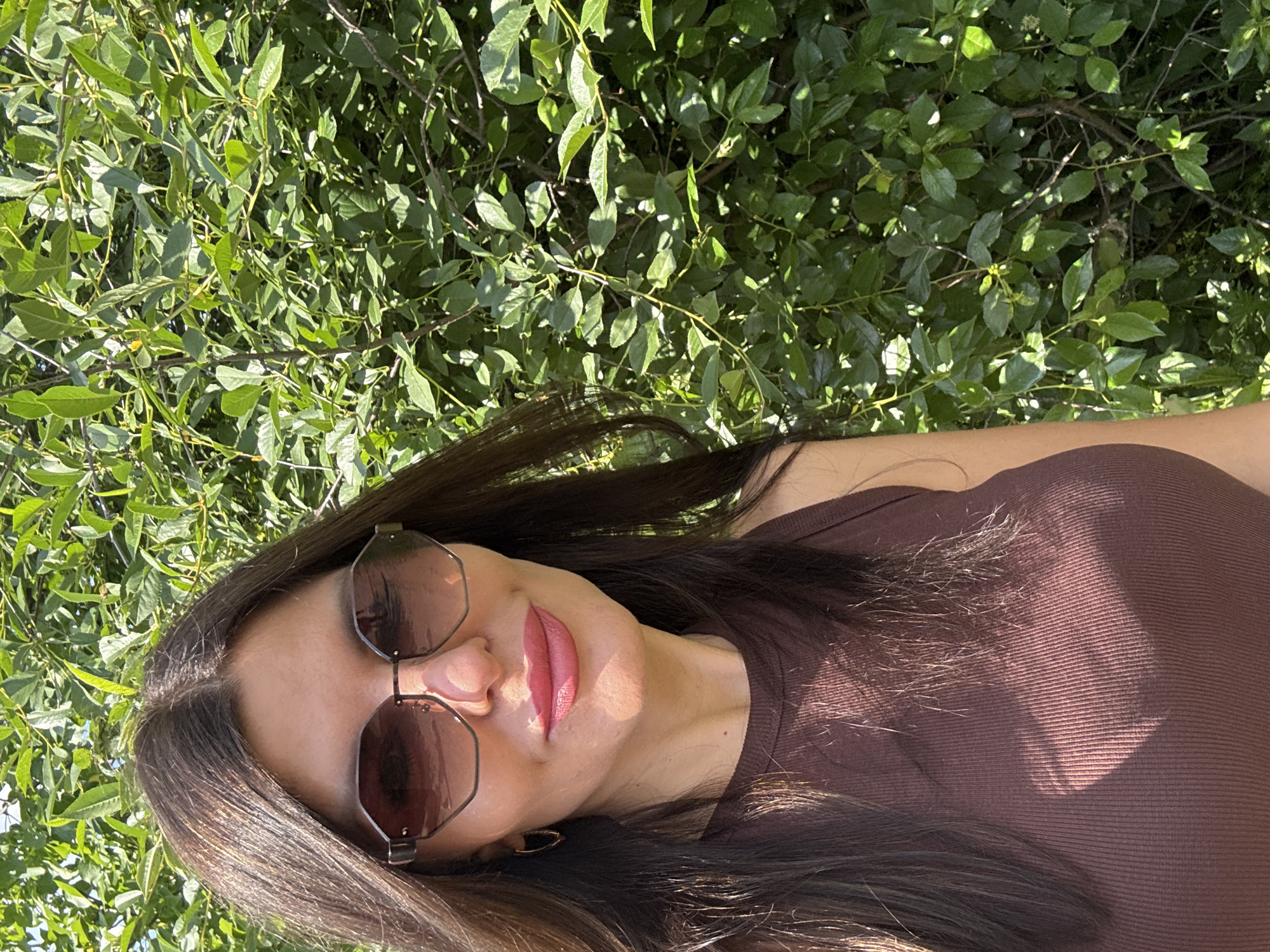 The image depicts a woman in a brown ribbed tank top and sunglasses, standing outdoors against a backdrop of dense green foliage. She has long, dark brown hair and is wearing pink lipstick. The sun casts shadows on her face and chest, suggesting it is daytime. The location is described as Chaikovsky, Russia, though no specific landmarks or indicators of this city are visible in the image. The scene is natural and appears to be taken in a park or garden.