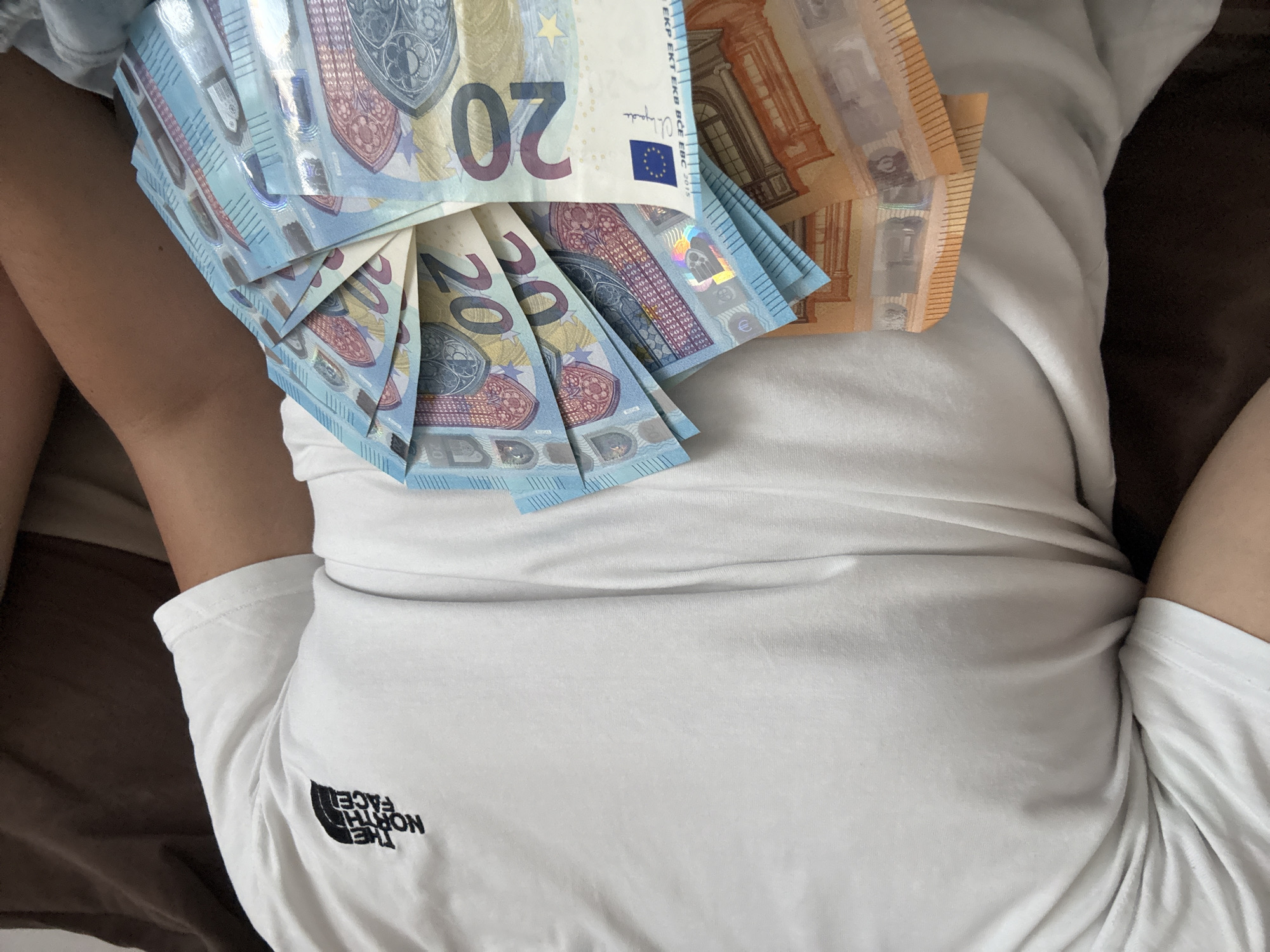 The image presents a close-up, high-angle view of a person lying down, partially visible from the chest to the upper thighs. The individual has a light skin tone and is wearing a plain white short-sleeved t-shirt. On the person's torso, a significant amount of Euro banknotes are arranged. A fan of twenty-euro notes, primarily light blue, is spread out in the center and to the left. To the right, a stack of orange fifty-euro notes is visible.
Visible text on the twenty-euro notes includes the denomination "20", "EURO", "EYPΩ", the acronyms for the European Central Bank in various languages ("ЕЦБ ЕКП ESB EZB EKT EKB BCE EBC 2015"), and a signature. A European flag symbol is also present. The fifty-euro notes display "50", "EURO", "EYPΩ", and the Central Bank acronyms ("ЕЦБ ЕКП ESB EZB EKT EKB BCE EBC 2017"). The white t-shirt features a black "THE NORTH FACE" logo, which appears inverted, consistent with the person lying on their back and the photo being taken from above. The background consists of dark brown fabric, likely bedding or a cushion, indicating an indoor, relaxed setting. The presence of Euros, within the context of Varna, Bulgaria, suggests the use of foreign currency. No specific activity beyond the display of money is discernable, and no emotions are visible.
