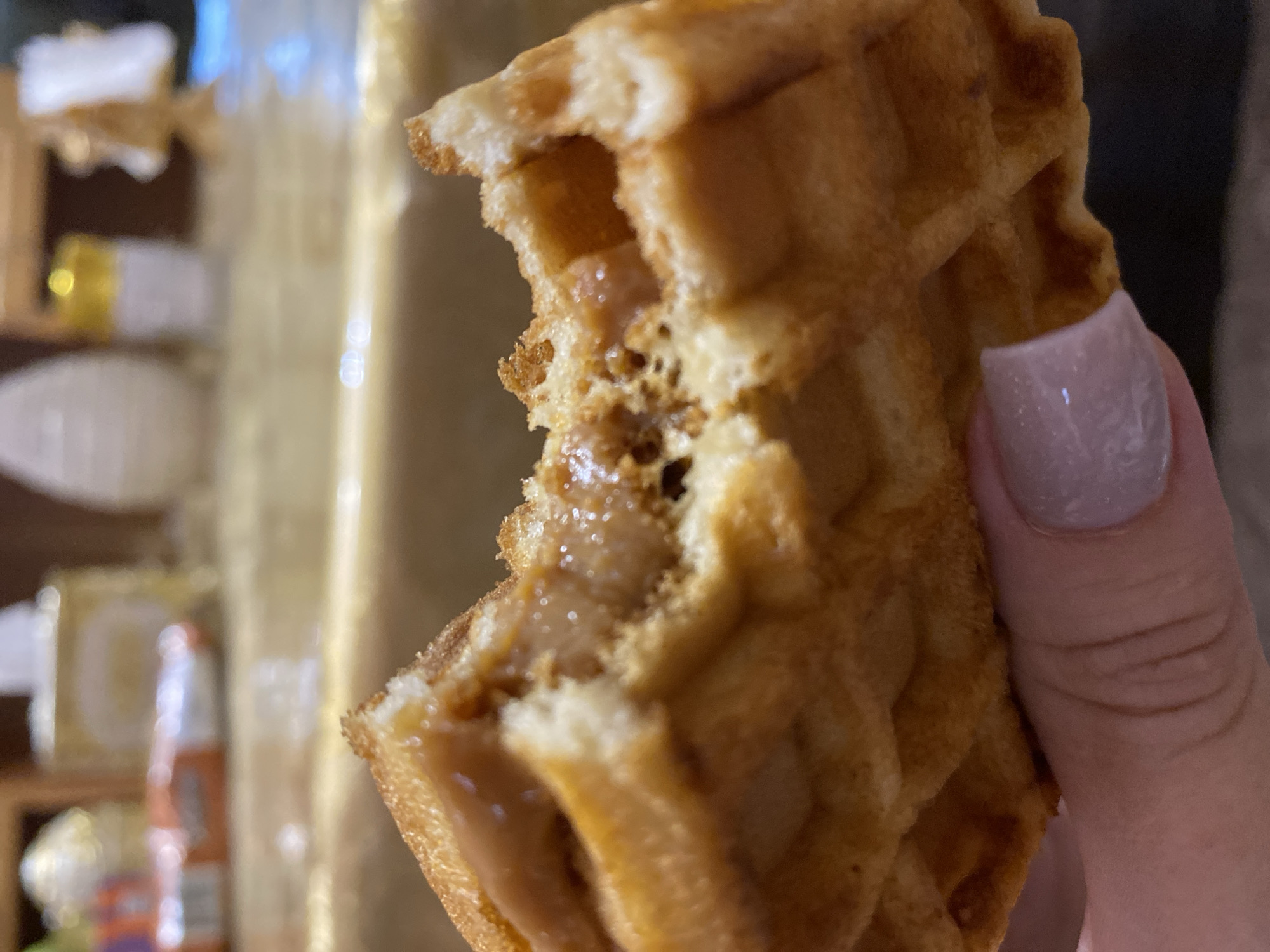 A close-up, shallow depth-of-field shot shows a person's thumb and index finger, with light pink nail polish, holding a bite-sized piece of a waffle with a caramel-like filling. The waffle is golden brown and has the characteristic grid pattern. The filling appears smooth and slightly glossy. In the background, a blurred kitchen counter with some out-of-focus items, possibly food packaging and dispensers, is visible. There is no identifiable location in Maykop, Russia.