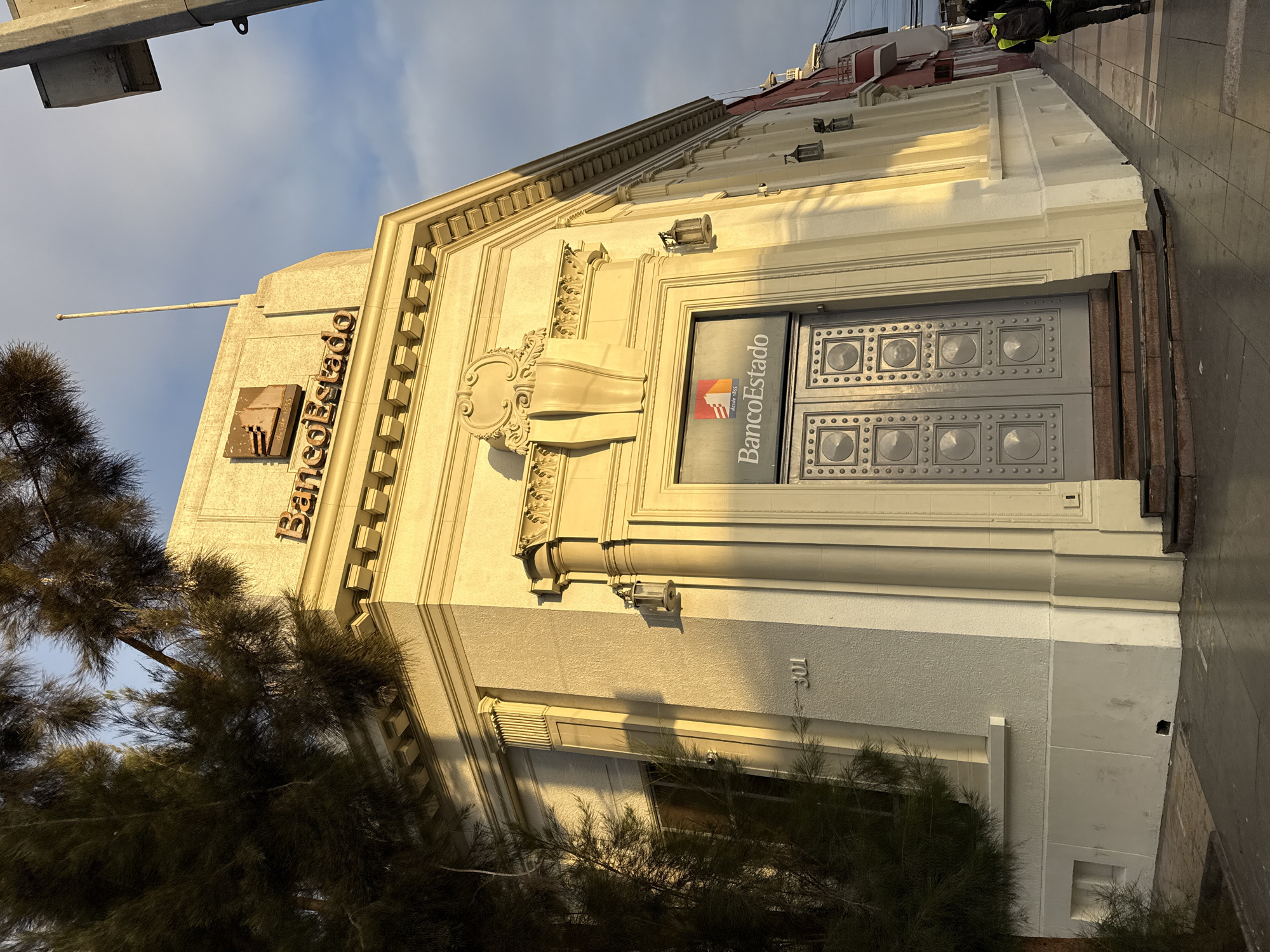 This image captures the BancoEstado building located in Iquique, Chile, on a clear day with dramatic lighting. The building, painted a pale yellow, exhibits classical architectural elements such as columns and decorative molding. The prominent entrance features a large, grey double door with a patterned design. Above the entrance, a glass panel displays the BancoEstado logo and the text "desde 1855". The building's facade is adorned with several wall-mounted lanterns. A tree with dark foliage partially obscures the left side of the building, and the number "301" is visible on the wall. The street in front of the bank appears wet, suggesting recent rain. A person wearing a bright yellow vest and a backpack is walking away from the camera on the sidewalk, to the right of the bank. The sky is a mix of blue and lighter hues, indicating either early morning or late afternoon light.