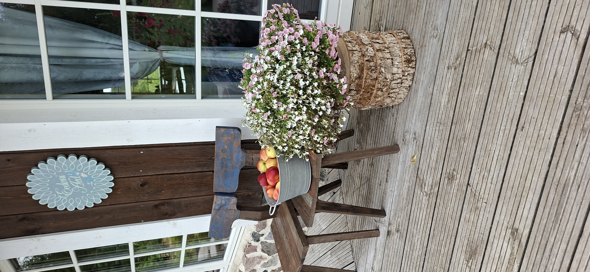 The image shows a rustic porch scene, likely in Pihali, Estonia. The porch is made of weathered wooden planks. To the left, a dark wooden door is mounted with a light blue, scalloped decorative sign featuring swirling lines and a small floral motif. Next to the door, a wooden chair with blue painted backrests is placed. In front of the chair, a galvanized metal bucket is filled with red and yellow apples. To the right of the chair, a large planter made of stacked wood slices overflows with small pink and white flowers. A second, lower wooden table is visible to the far left of the image, also made of weathered wood. Behind the scene, a white-framed window with sheer blue curtains is visible, offering a glimpse of greenery outside. The overall atmosphere is peaceful and suggestive of a rural or garden setting.