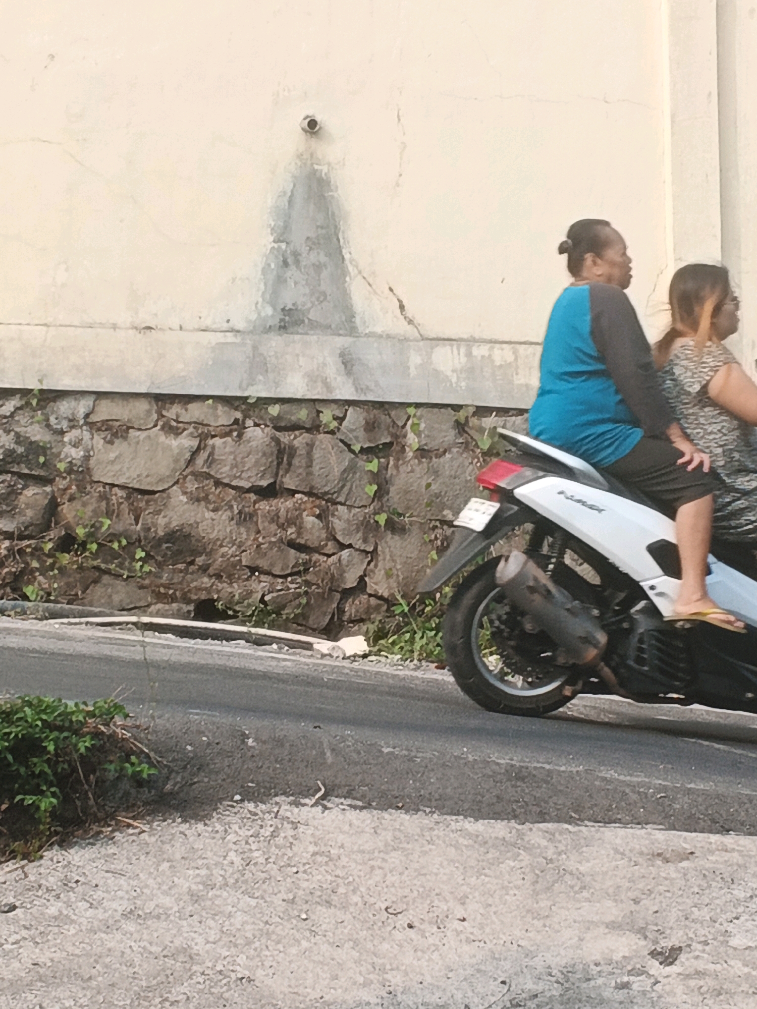 Two women are riding a white scooter on a street in Keji, Indonesia. The woman in front is wearing a blue long-sleeved shirt and black shorts. The woman in the back is wearing a patterned top. The scooter is white with black accents and has a black exhaust pipe. The background consists of a stone wall and a beige wall with some water stains. The ground is paved with asphalt and gravel. There are some green plants growing along the stone wall and on the side of the road. The lighting suggests it is daytime.