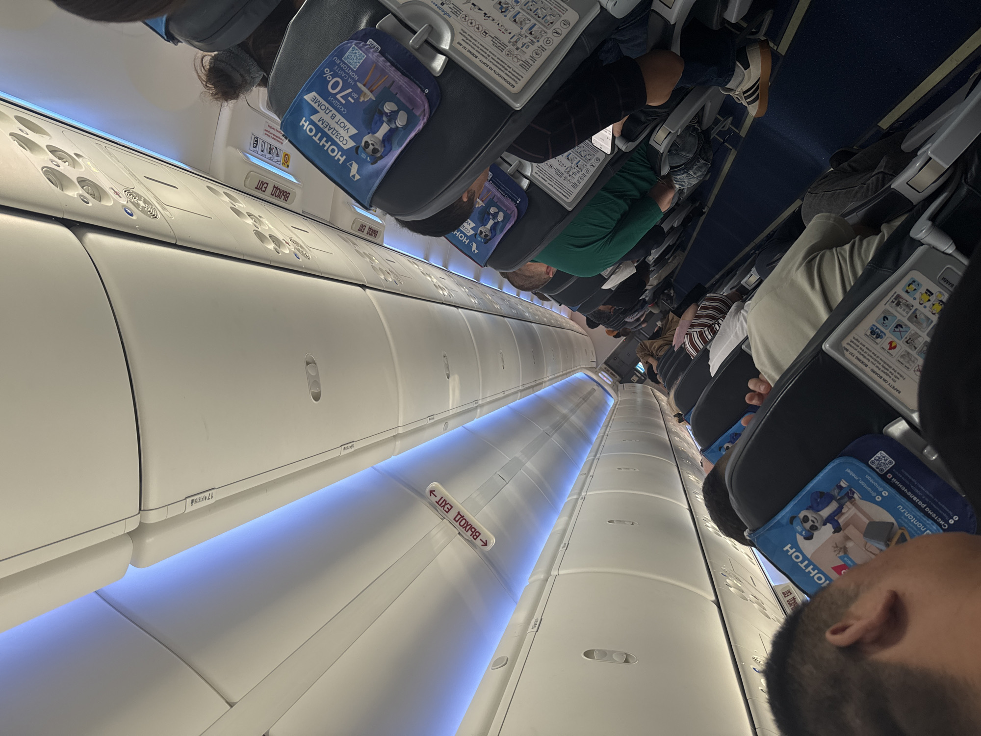 The image shows the interior of an airplane cabin, likely during a flight, with passengers seated. The perspective is looking down the aisle from the front of the plane. Overhead compartments and lighting are visible, with blue LED lights illuminating the ceiling. Several "EXIT" signs are present, written in both Russian and English. Some passengers have their heads down, possibly resting or using electronic devices. Seatback pockets contain promotional materials, including one with a panda mascot and text advertising discounts. The overall environment suggests a commercial flight in progress, with typical airplane seating and amenities. The question about the location in Arnavutköy, Turkey is not directly answerable from the visual information provided.