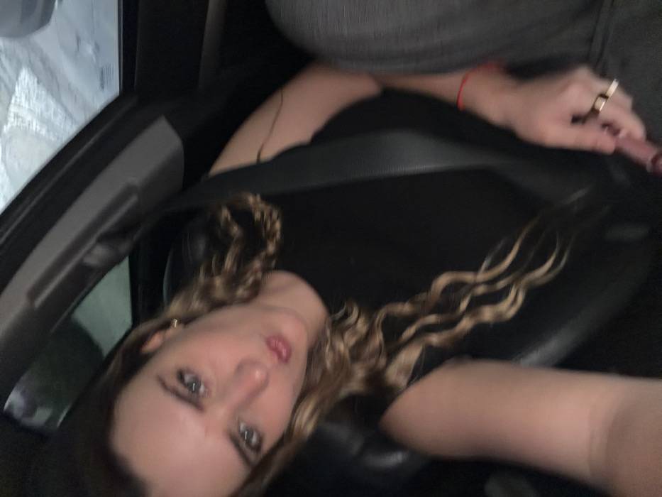The scene is situated inside a vehicle, understood to be in San Bernardino Tlaxcalancingo, Mexico, at night or in a dimly lit environment. A young adult female, with light brown or blonde wavy hair and blue eyes, is in the passenger seat. She wears a black sleeveless top and is secured by a black seatbelt across her chest. Her head is tilted, and she looks towards the camera with pursed lips. Her left hand, wearing a red string bracelet on the wrist and a gold ring on the ring finger, holds a pink cylindrical object. The background beyond the vehicle's interior shows blurred lights, possibly reflections on a wet windshield.