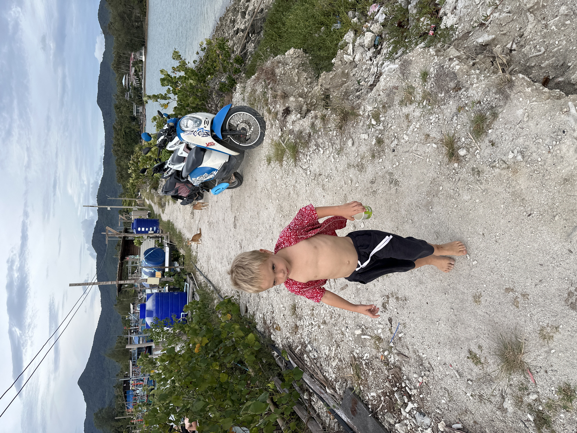 A young Caucasian boy with blond hair walks barefoot on a dirt path. He is wearing a red shirt open over his bare chest and black shorts. He holds a small white cup in his right hand. The path is lined with green bushes on the left and a body of water on the right. In the background, there are several blue water tanks, boats, and a small orange cat sitting on the path. Further in the distance, mountains and palm trees are visible under a cloudy sky. Several scooters are parked on the right side of the path. The scene appears to be in a coastal area of Ko Pha-ngan District, Thailand.