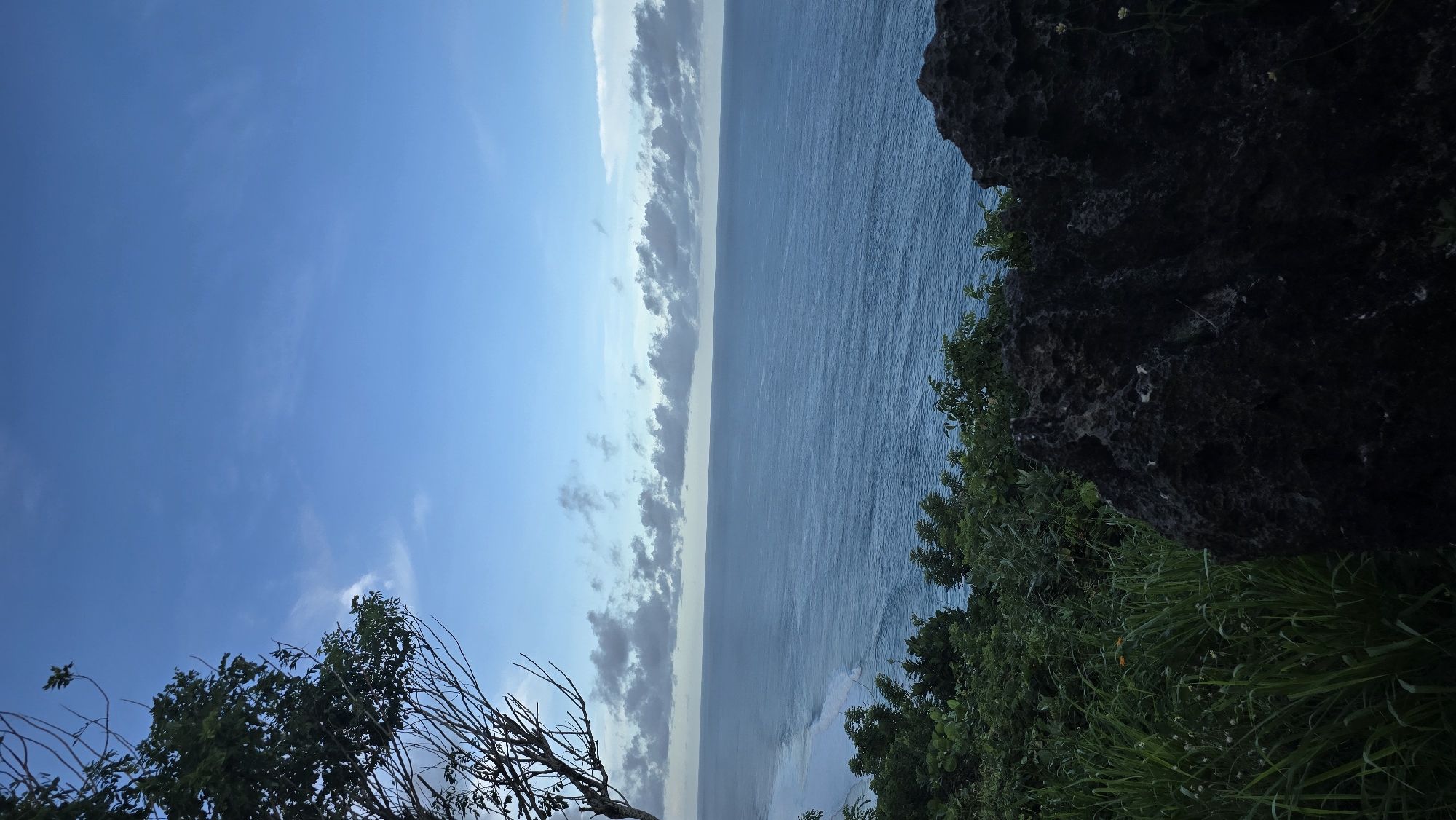 The image captures a coastal scene likely in Kuta Selatan, Indonesia. A blue sky with scattered clouds covers the left portion of the image. The ocean occupies the central space, displaying a variety of blue tones. A line of clouds meets the ocean. A rocky cliff face with green vegetation frames the right side of the picture. The lower left corner features a cluster of trees and foliage.
