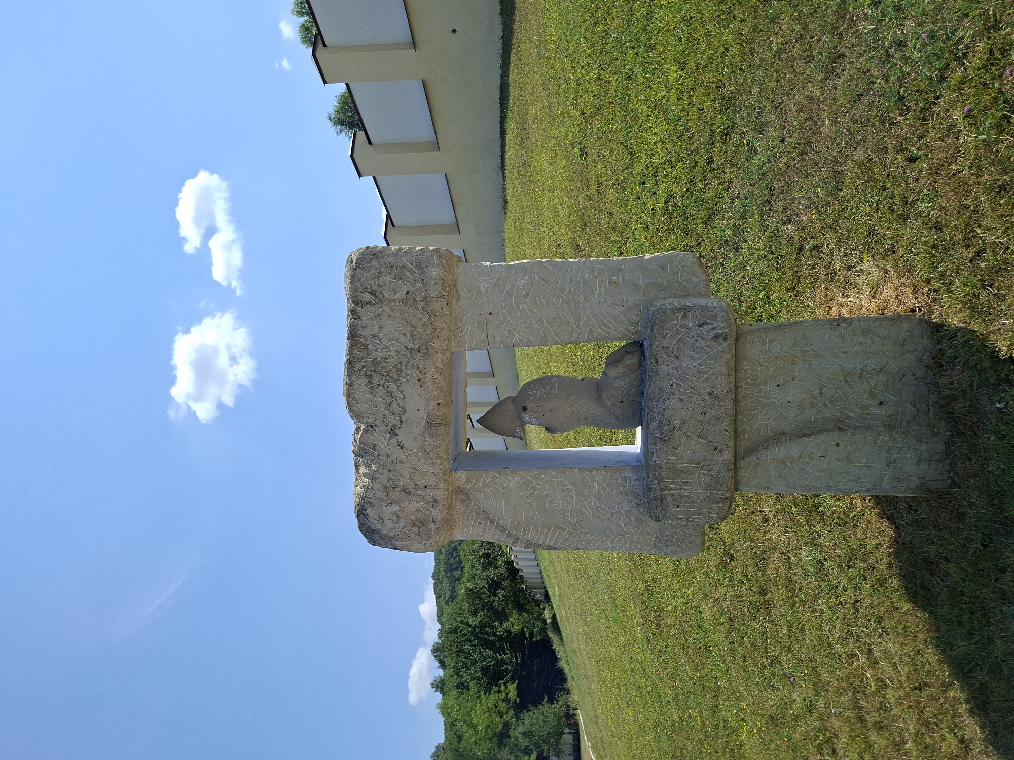 An outdoor, eye-level, medium shot shows a stone sculpture with a figure inside, located in the city of Condrița, Moldova. The sculpture is made of light-colored, rough-hewn stone, with visible scratches and carvings on its surface. The central part of the sculpture frames a smaller, abstract figure that appears to be kneeling or seated, wearing a hooded garment. This figure is also made of stone. The sculpture stands on a grassy knoll under a bright blue sky with scattered white clouds. Behind the sculpture, a light yellow wall with dark-topped vertical pillars stretches across the middle ground. The wall is set against a backdrop of lush green trees on a hillside to the left and further down the slope to the right. The grass in the foreground is a mix of green and dry patches, with some small yellow and pink wildflowers. A dark shadow is cast by the sculpture on the grass to its left. The overall scene depicts a sunny day in a rural or park-like setting.