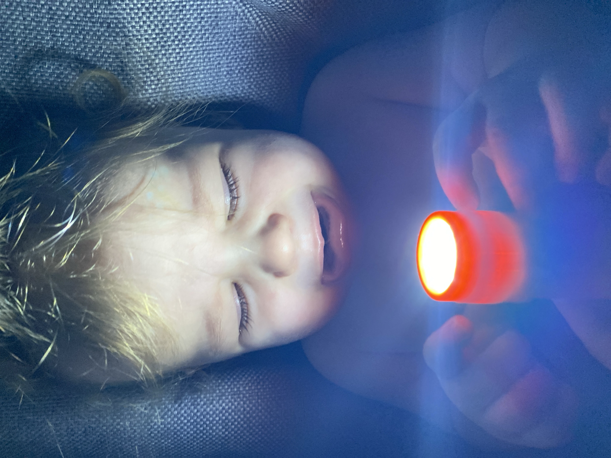 A close-up, low-angle shot shows a child lying in what appears to be a dark, textured surface, possibly a stroller or seat. The child has fair skin, blonde hair, and is looking up with their mouth slightly open and eyes nearly closed. In their hand, they hold a small flashlight with a bright orange-tinted beam illuminating their face and hand. The background is blurred due to the low light and shallow depth of field. The overall color tone is cool, with blue hues dominating the shadows. The location is unknown.