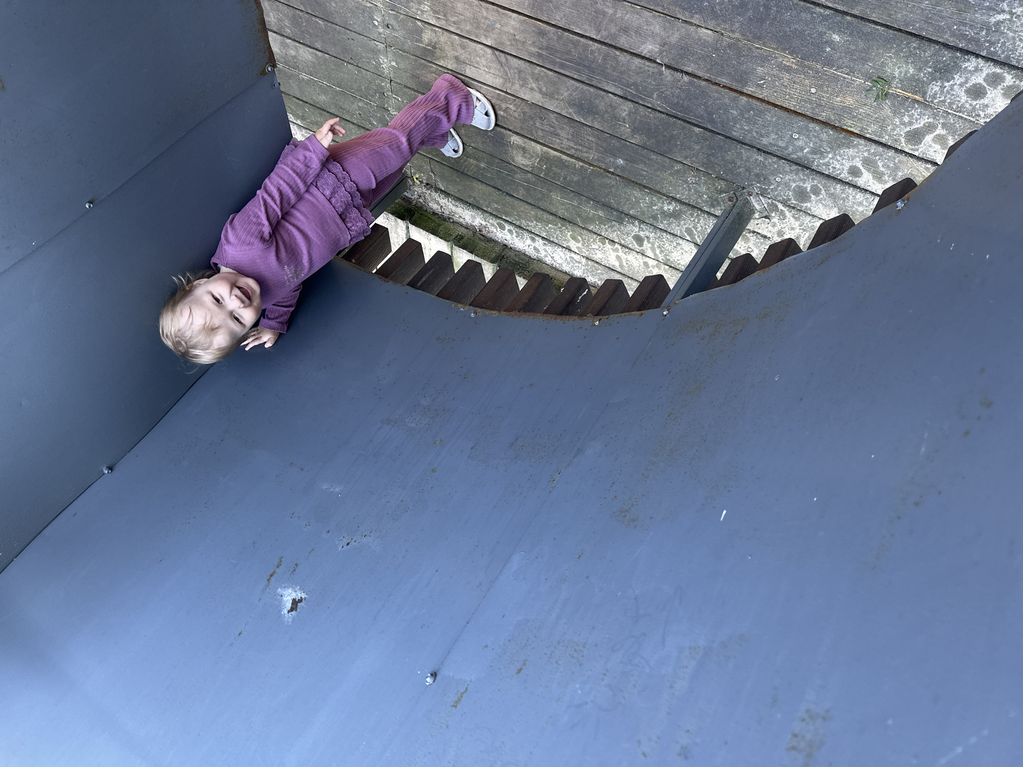 A full-length, low-angle shot captures a smiling young child, likely a toddler, lying on an inclined dark blue-grey metal surface. The child has light blonde hair and is dressed in a matching purple long-sleeved top and pants, with light-colored sandals on their feet. Their head is turned towards the camera, revealing a wide, joyful smile.
The metal surface, possibly a ramp or part of a slide, shows signs of wear such as minor scratches, dirt, and small rust spots. Behind and above the child is a wooden platform or deck made of weathered planks. These planks exhibit discolored patches, possibly from moss or moisture, and general wear, indicating an outdoor environment. Below the wooden platform and the edge of the metal structure, dark wooden slats or supports are visible, forming a barrier or part of the structure's foundation.
The overall scene suggests a playful moment at an outdoor playground or recreational area during the daytime, with soft, indirect lighting. The child's happy expression indicates enjoyment. The location context for this scene is Nizhny Novgorod, Russia. No text is visible in the image.